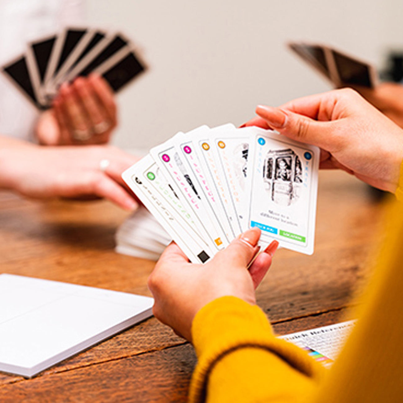 A close-up of hands playing a card game over a wooden table, holding a fan of illustrated cards. The scene shows multiple people interacting in a casual indoor setting.