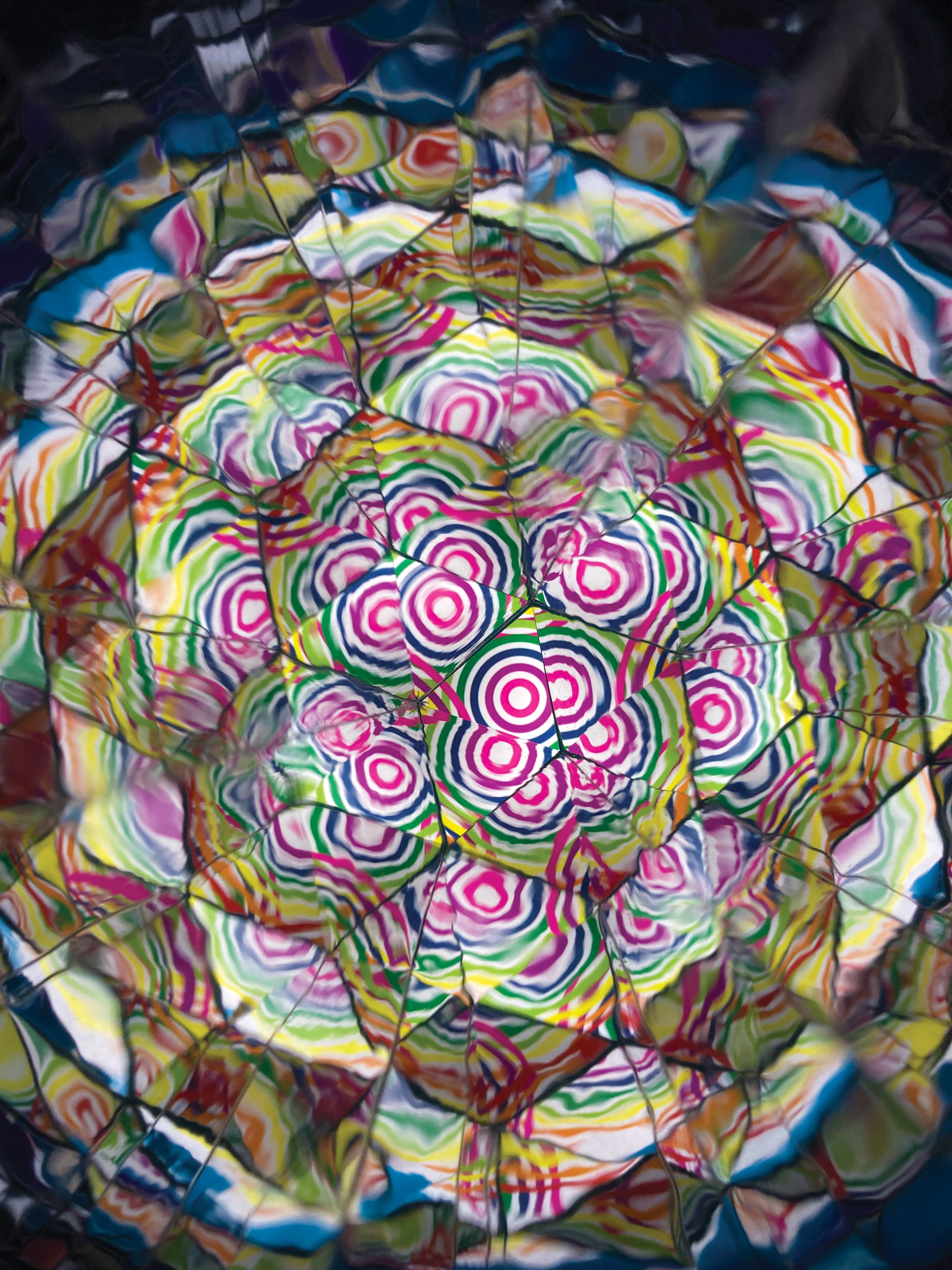 A highly colourful abstract circular artwork with concentric floral or spiral motifs in pink, green, yellow, blue, and red, overlaid with dark web-like lines.