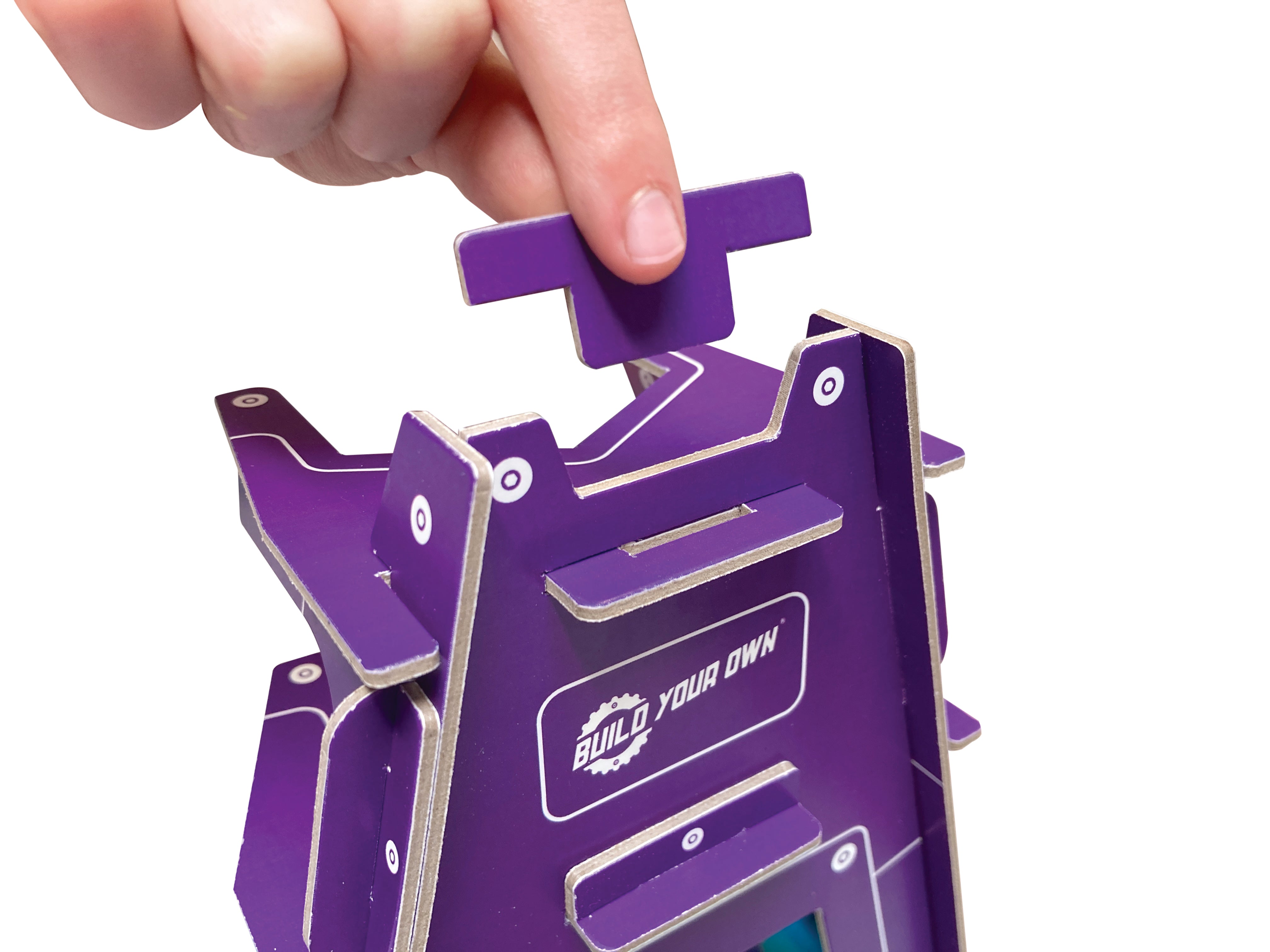 A close-up of a hand assembling a purple cardboard model kit, pressing a tab onto a built structure with the words 'BUILD YOUR OWN' printed on the side, set against a plain white background.