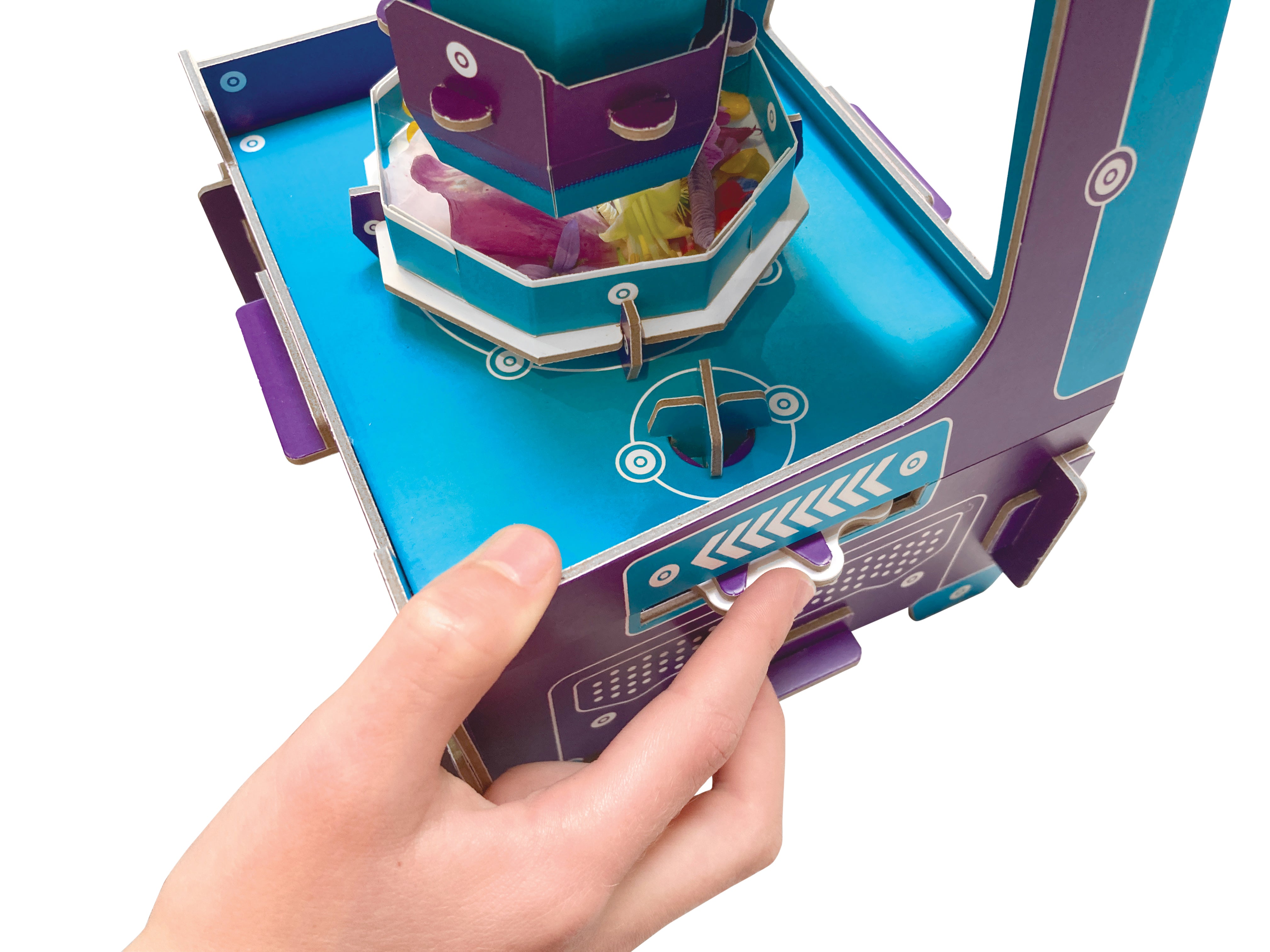 A close-up of a hand operating a colourful paper/cardboard toy machine with a blue base, purple side panels, and a central tower structure, photographed on a white background.