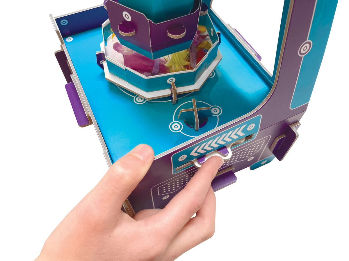 A close-up of a hand operating a colourful paper/cardboard toy machine with a blue base, purple side panels, and a central tower structure, photographed on a white background.