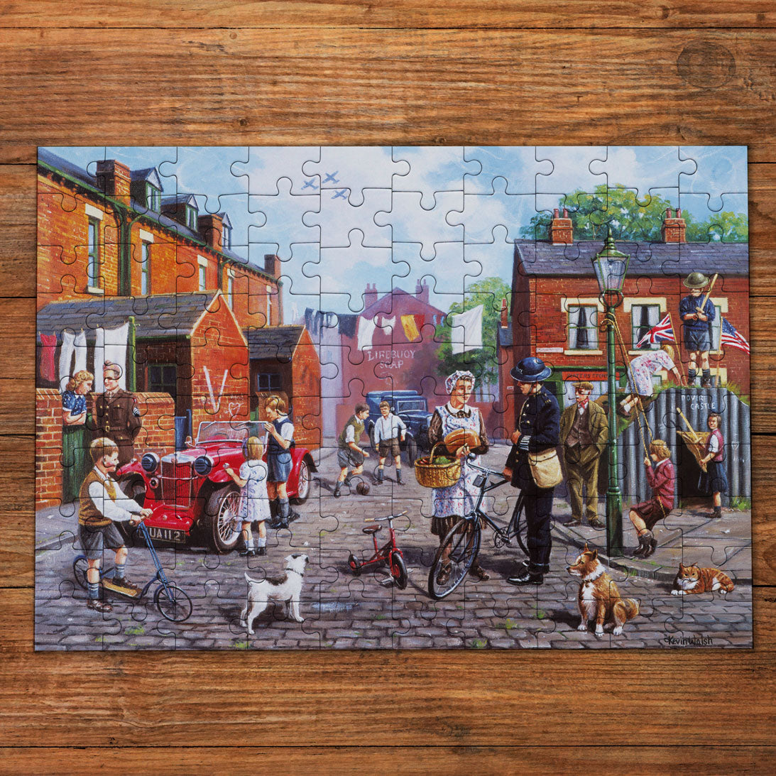 A completed jigsaw puzzle depicting a colorful, busy street scene with people, children, bicycles, a red car, and brick houses, displayed on a rustic wooden surface.