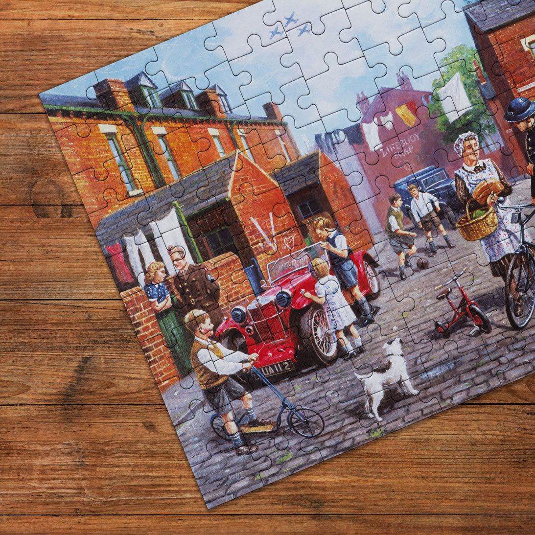 A completed jigsaw puzzle showing a colourful vintage street scene with people, a red car, bicycles, and a dog, placed on a wooden tabletop.