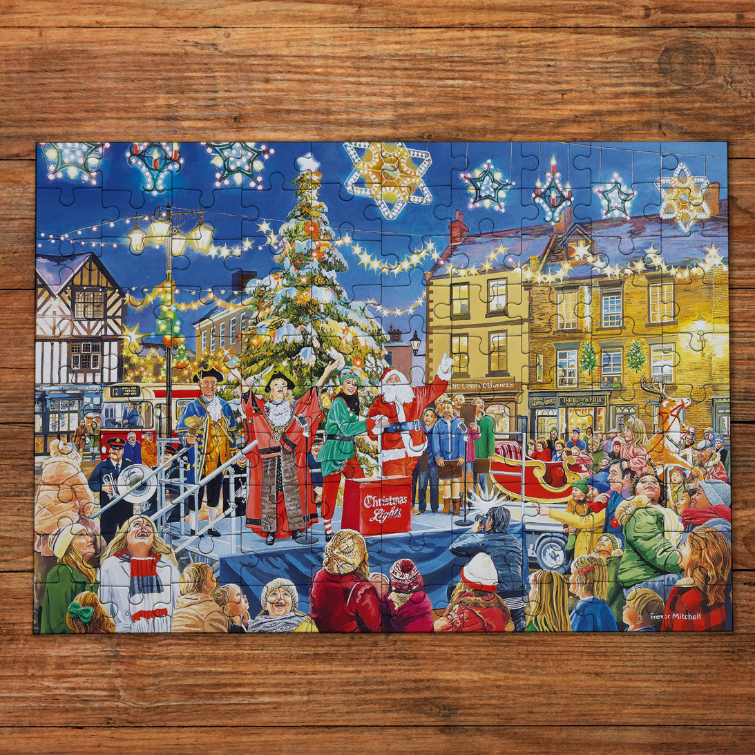 A completed jigsaw puzzle featuring a festive Christmas village scene with a decorated tree, performers, buildings, and crowds, placed on a wooden surface.