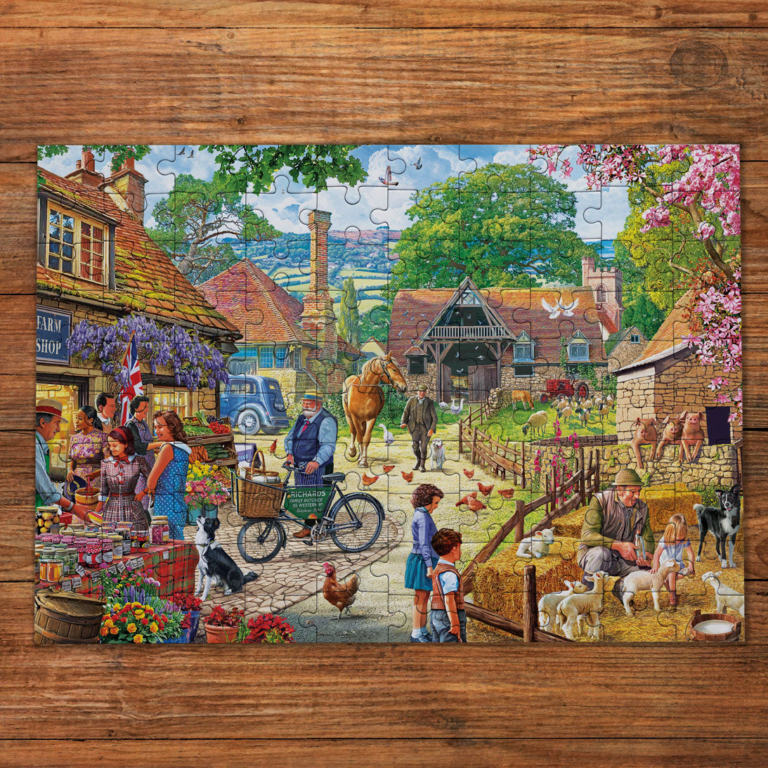 A completed jigsaw puzzle depicting a colourful rural village scene, placed on a rustic wooden surface. The scene includes people at a market, a horse and cart, cows, sheep, flowers, and farm animals.