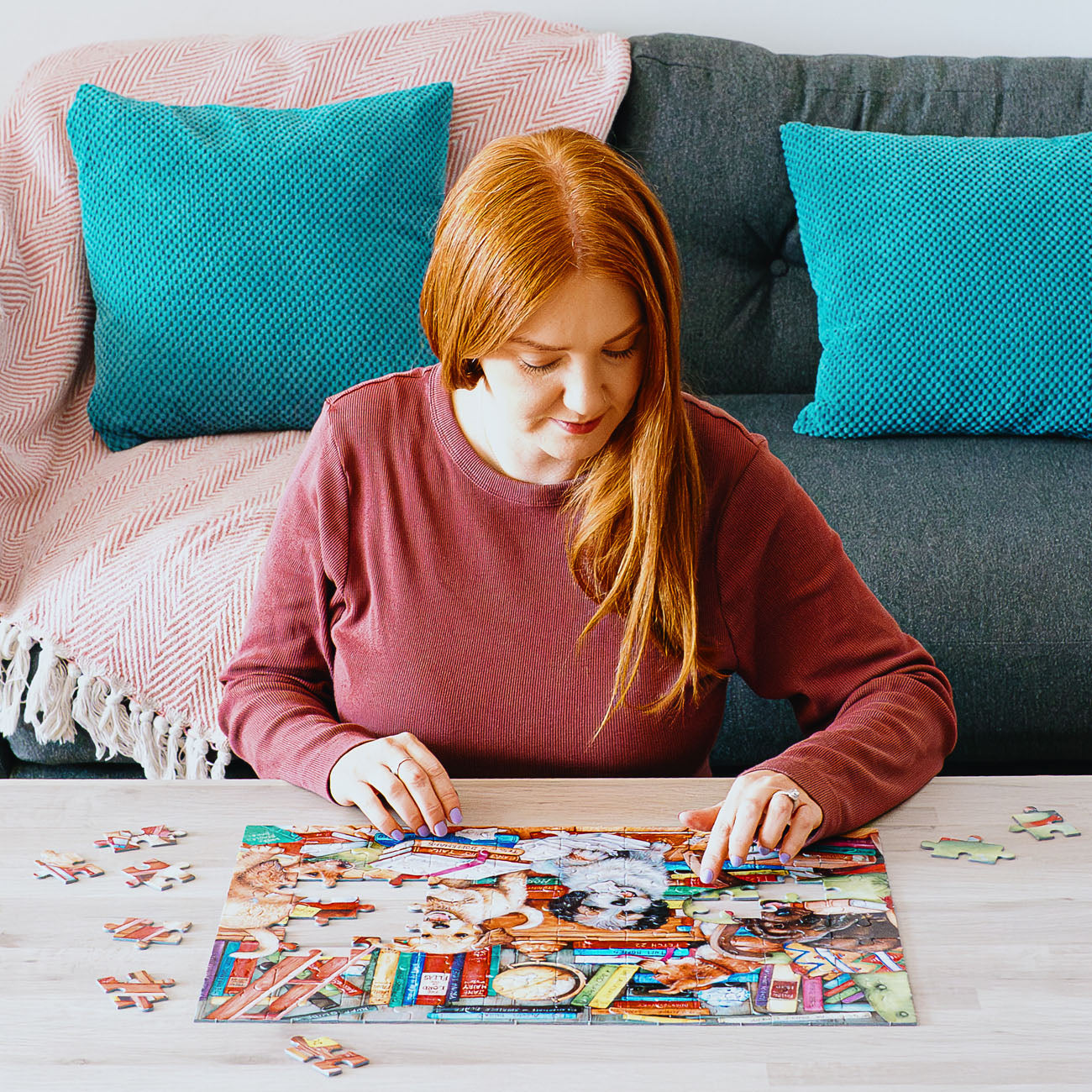 Woman with red hair sitting on a couch, working on a puzzle at a table.