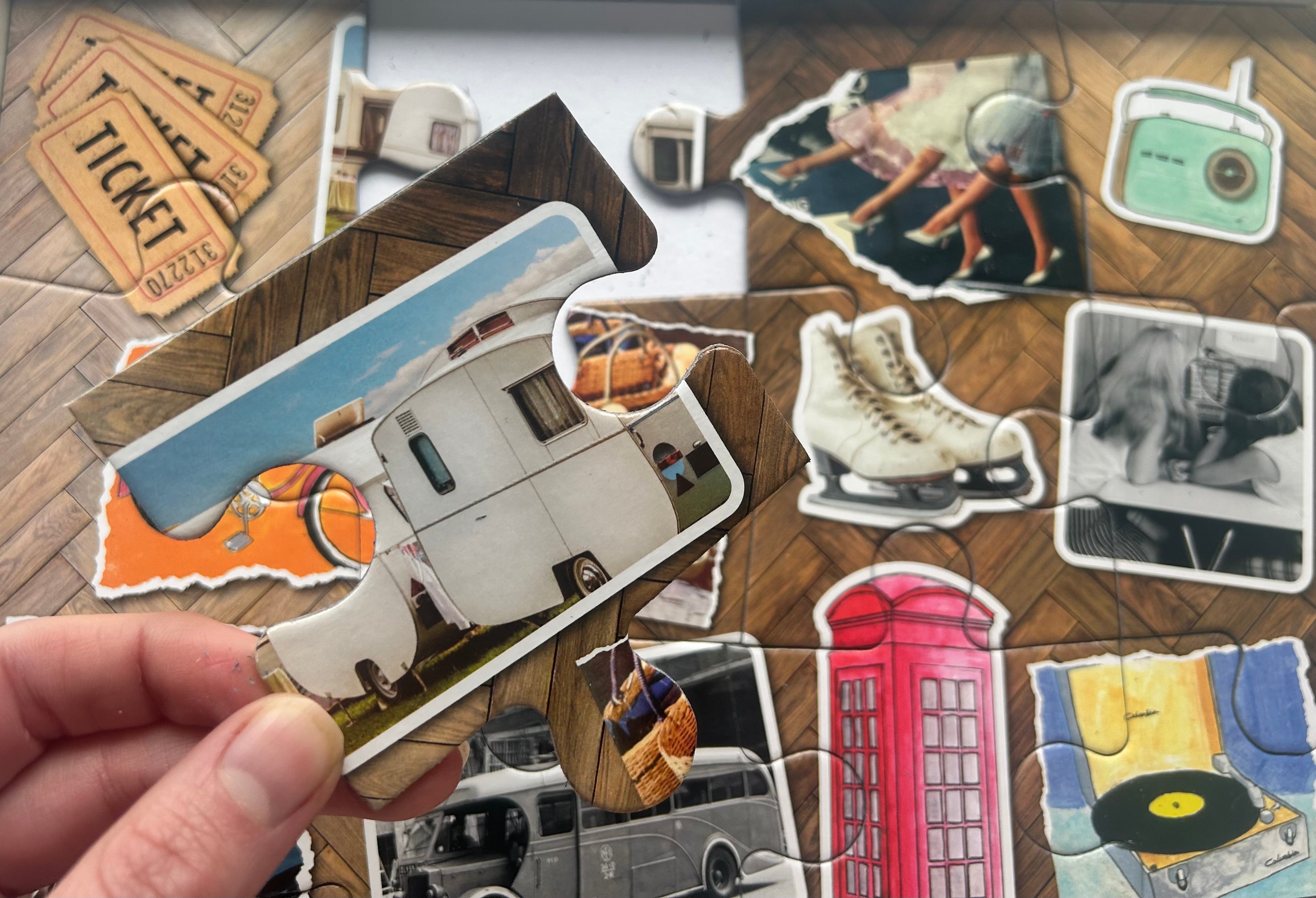 A hand holds a jigsaw puzzle piece shaped like a vintage trailer over a larger collage of matching vintage-themed puzzle pieces on a wood floor. Other visible pieces include a ticket, camera, ice skates, telephone booth, record player, and other retro objects.