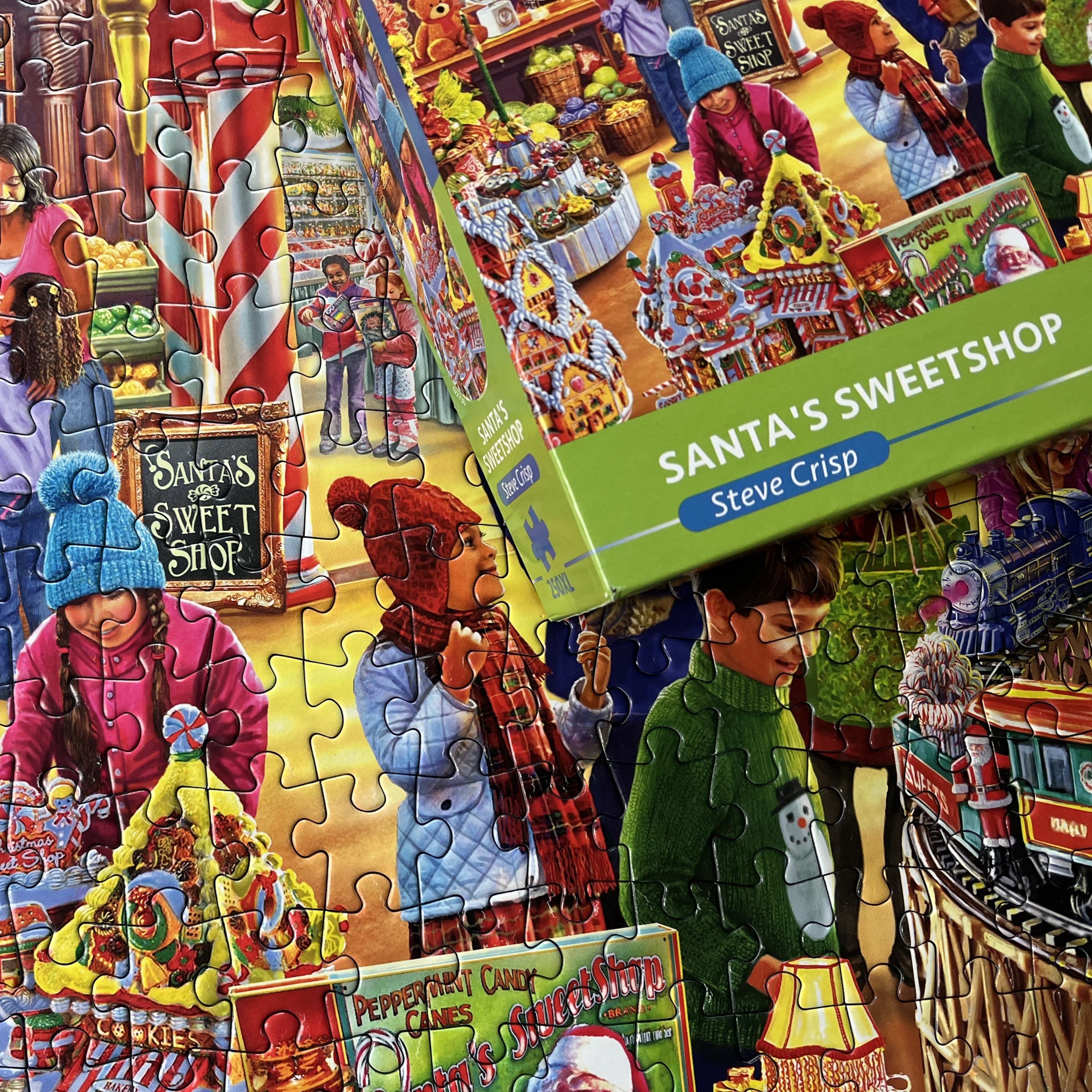 A completed jigsaw puzzle and its green box titled 'Santa's Sweetshop' by Steve Crisp, showing a colourful illustrated scene of children in winter clothing at a candy shop.