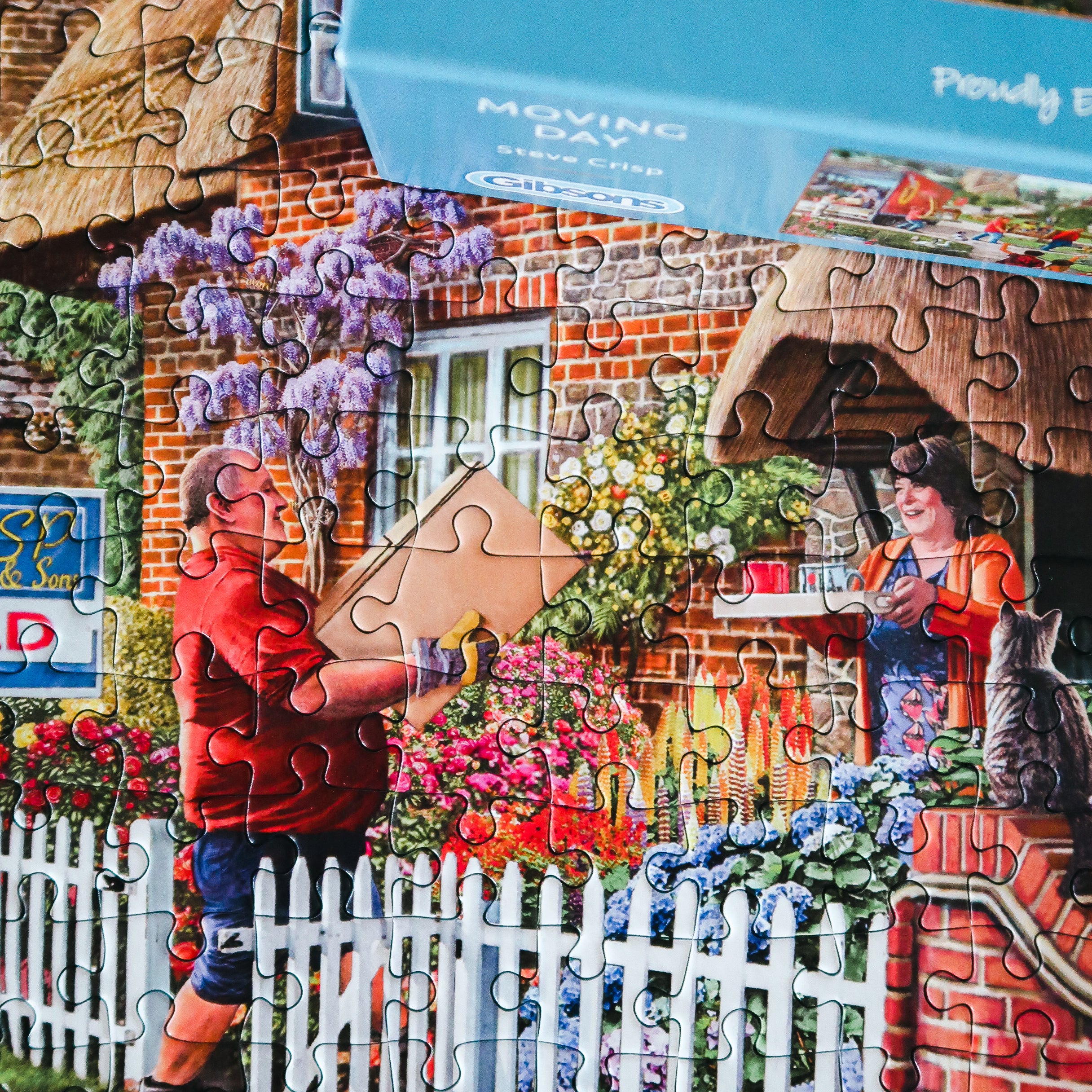 A completed jigsaw puzzle showing a colourful village scene with a brick house, white picket fence, flowers, and two people carrying a cardboard box, with a blue puzzle box header visible at the top.