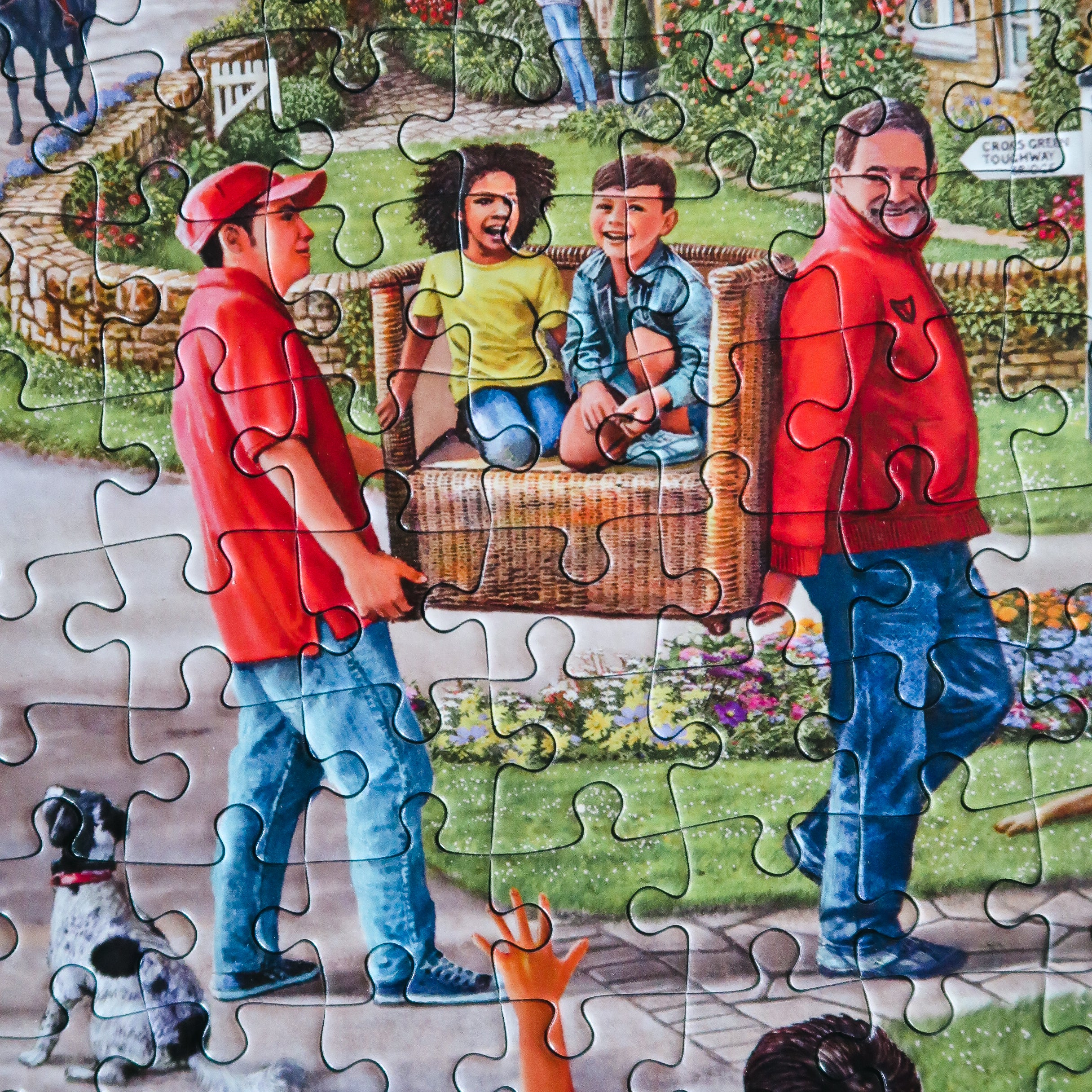 A completed jigsaw puzzle showing a colourful painted scene of two men carrying a wicker chair with two children inside, set in a suburban yard with a dog and flowers.