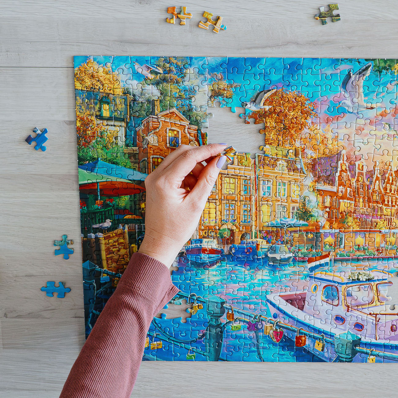 Person assembling a puzzle of a colourful cityscape with boats on a wooden table.