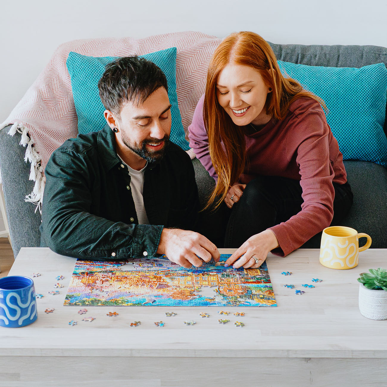 A man and a woman are sitting on a grey couch, smiling and assembling a colourful jigsaw puzzle on a white table. Blue mugs, a small potted succulent, and teal pillows are visible in the scene.