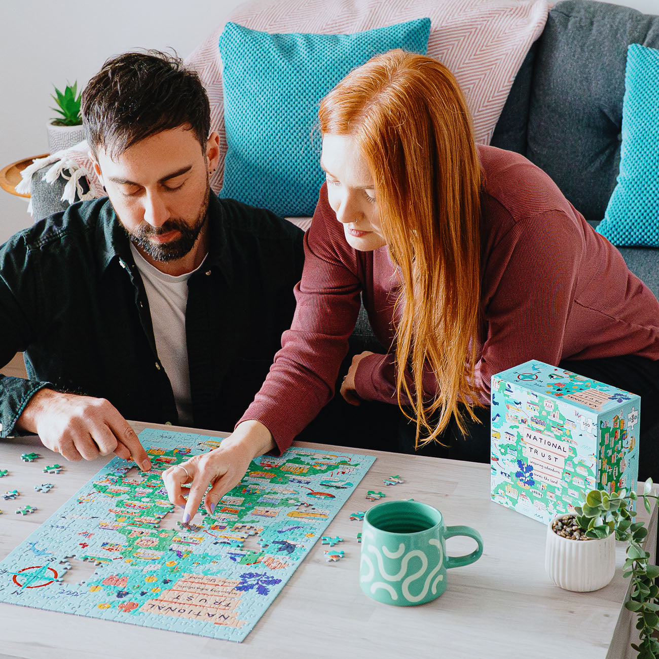 Two people working on a puzzle together with a mug and box in a cozy living room setting.