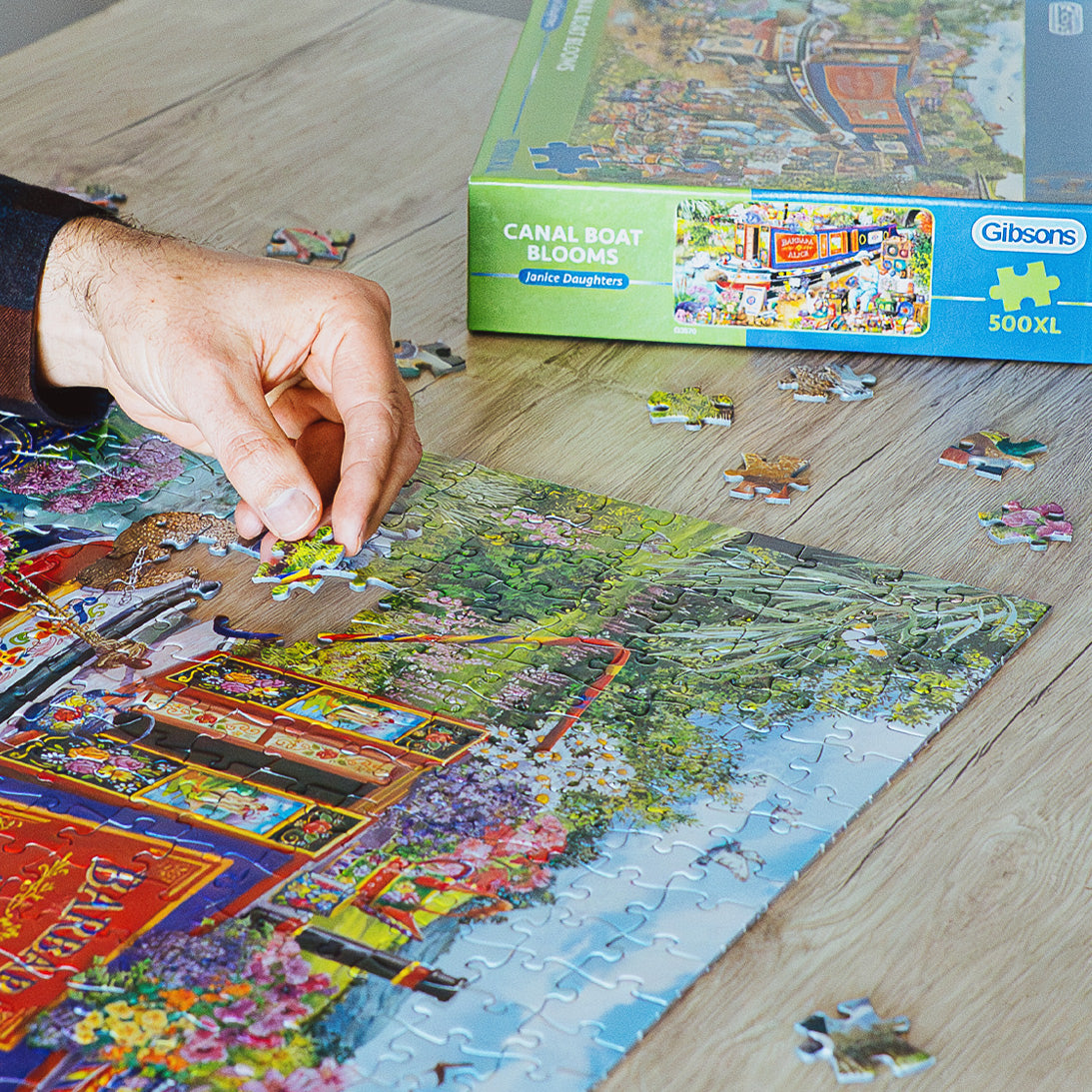 Person assembling a puzzle with a completed section, Gibsons puzzle box in the background