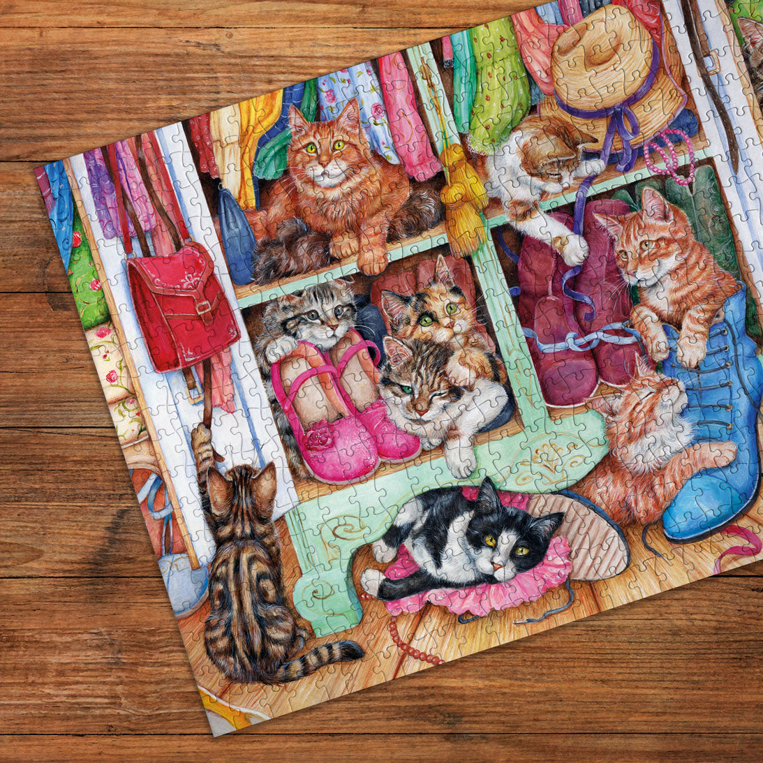 A completed jigsaw puzzle featuring a colorful cat-themed closet scene, photographed at an angle on a wooden tabletop. The puzzle shows several cats, pink shoes, a red handbag, colorful garments, and a blue boot, with the puzzle pieces still visible around the edges.