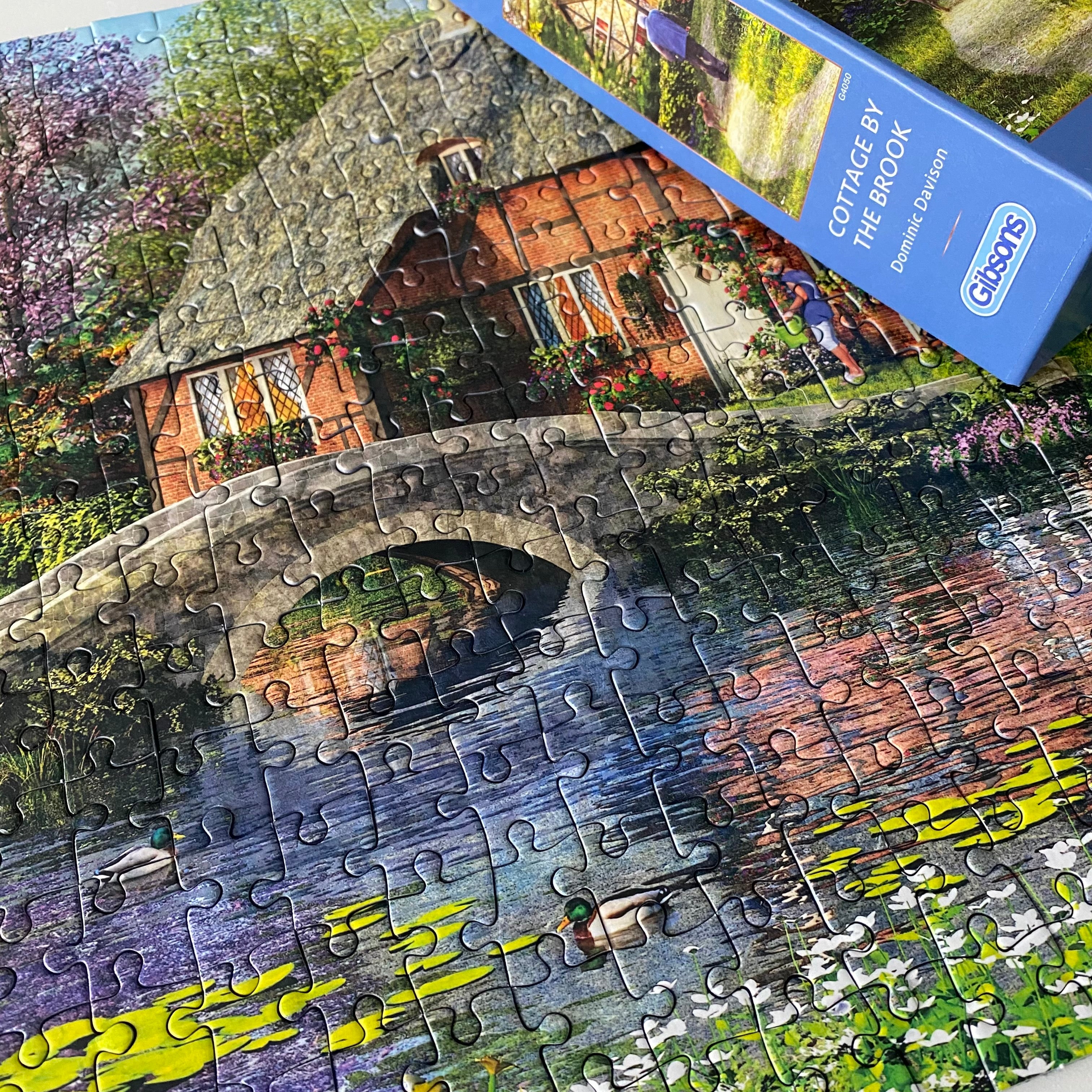 A completed jigsaw puzzle showing a picturesque cottage by a stone bridge over water with ducks, alongside the blue box packaging labelled 'Cottage by the Brook'.