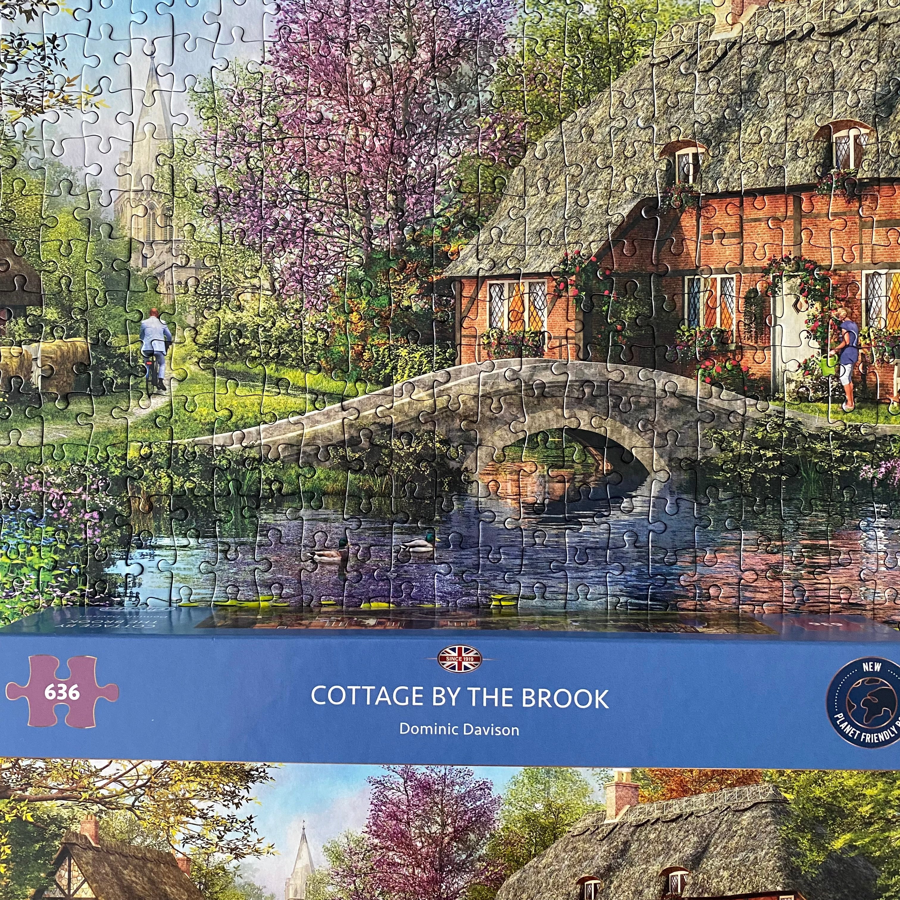 A completed jigsaw puzzle showing a picturesque cottage by a brook with a stone bridge, water reflection, and a stone wall in the background. The bottom of the puzzle features a blue band with the text 'COTTAGE BY THE BROOK', 'Dominic Davison', and puzzle details including '636 pieces'.