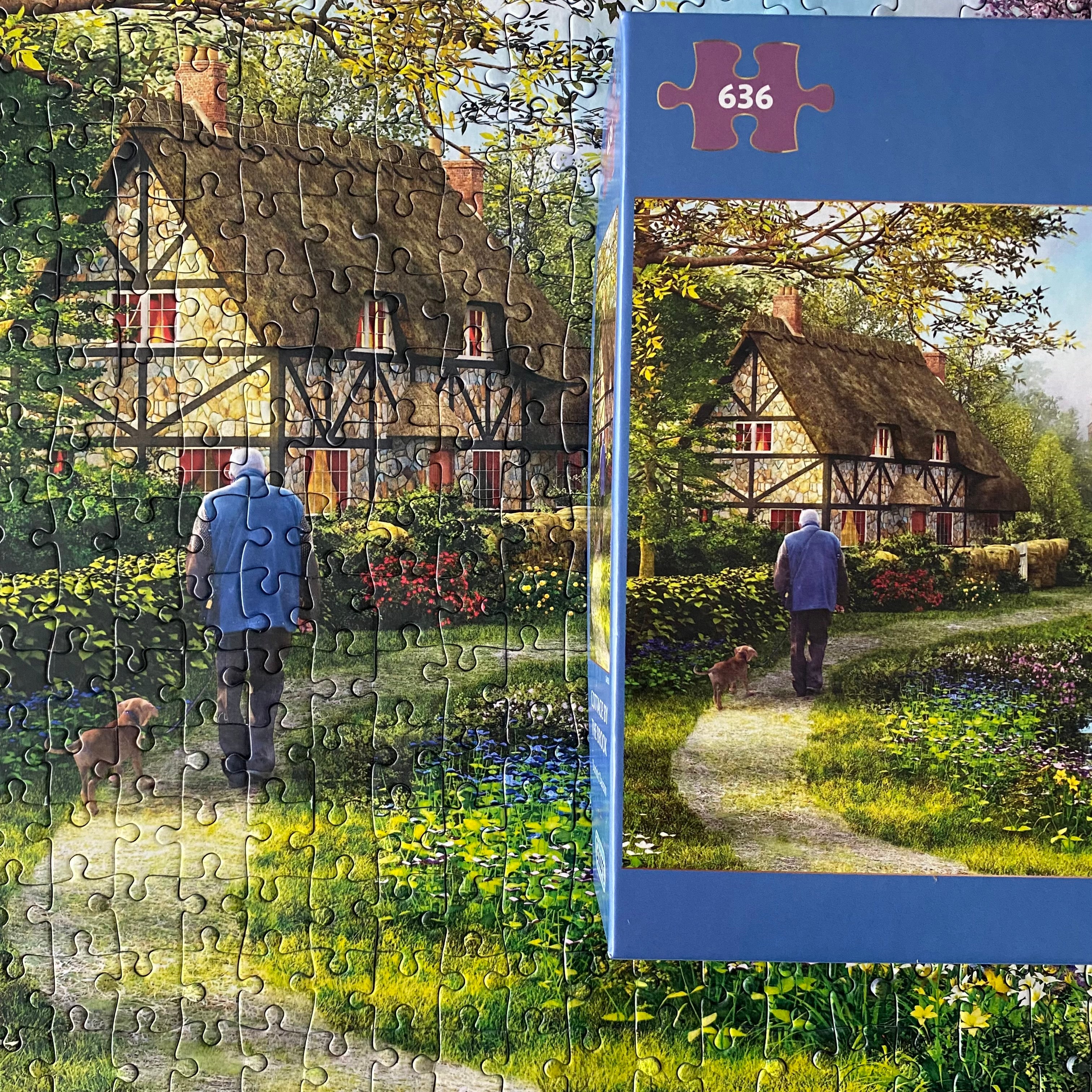A completed jigsaw puzzle showing a picturesque thatched cottage and a man walking with a dog in a flower-lined path, alongside its blue packaging box labeled '636'.