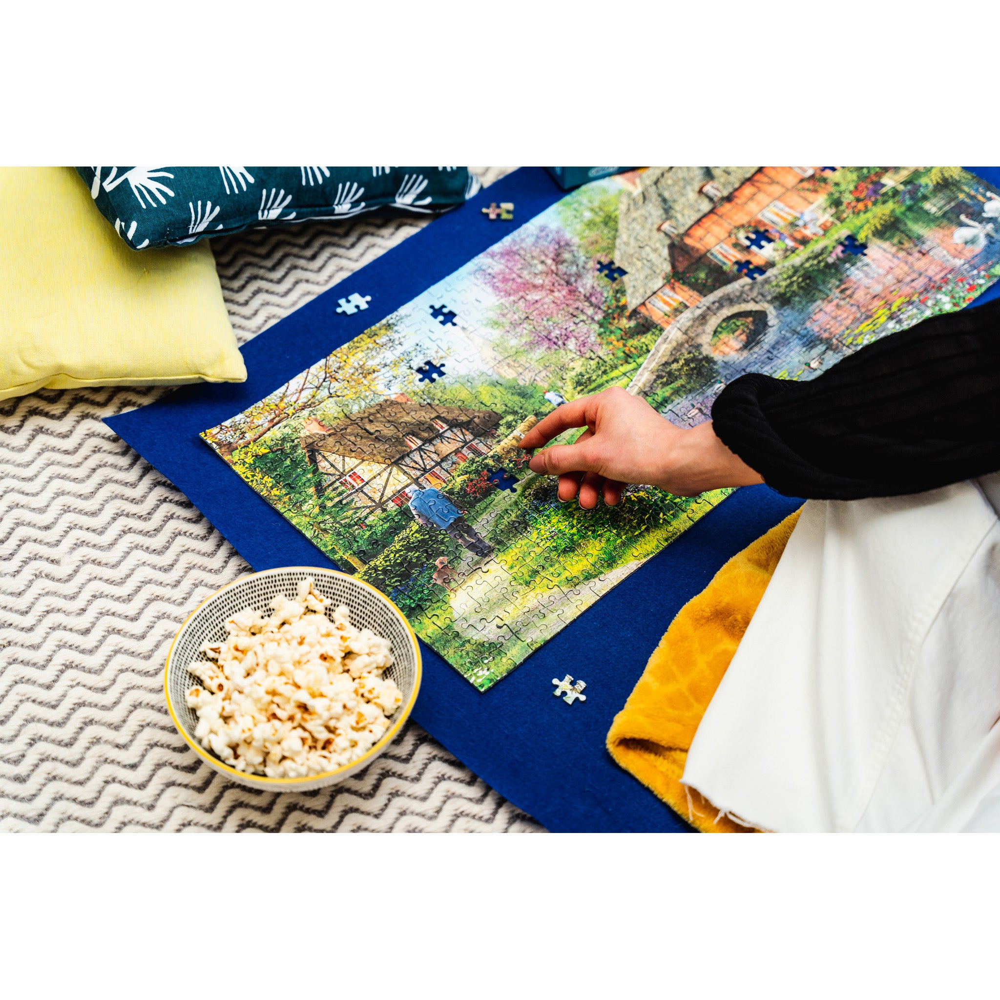 A person's hand placing a small jigsaw puzzle piece onto a colourful landscape jigsaw puzzle spread out on a blue puzzle roll, with pillows and a bowl of popcorn nearby on a patterned rug.