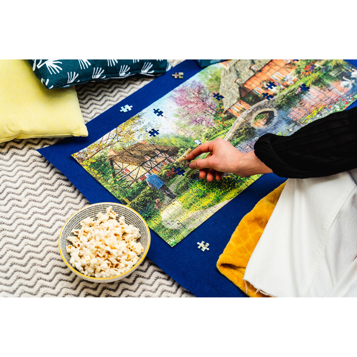 A person's hand placing a small jigsaw puzzle piece onto a colourful landscape jigsaw puzzle spread out on a blue puzzle roll, with pillows and a bowl of popcorn nearby on a patterned rug.