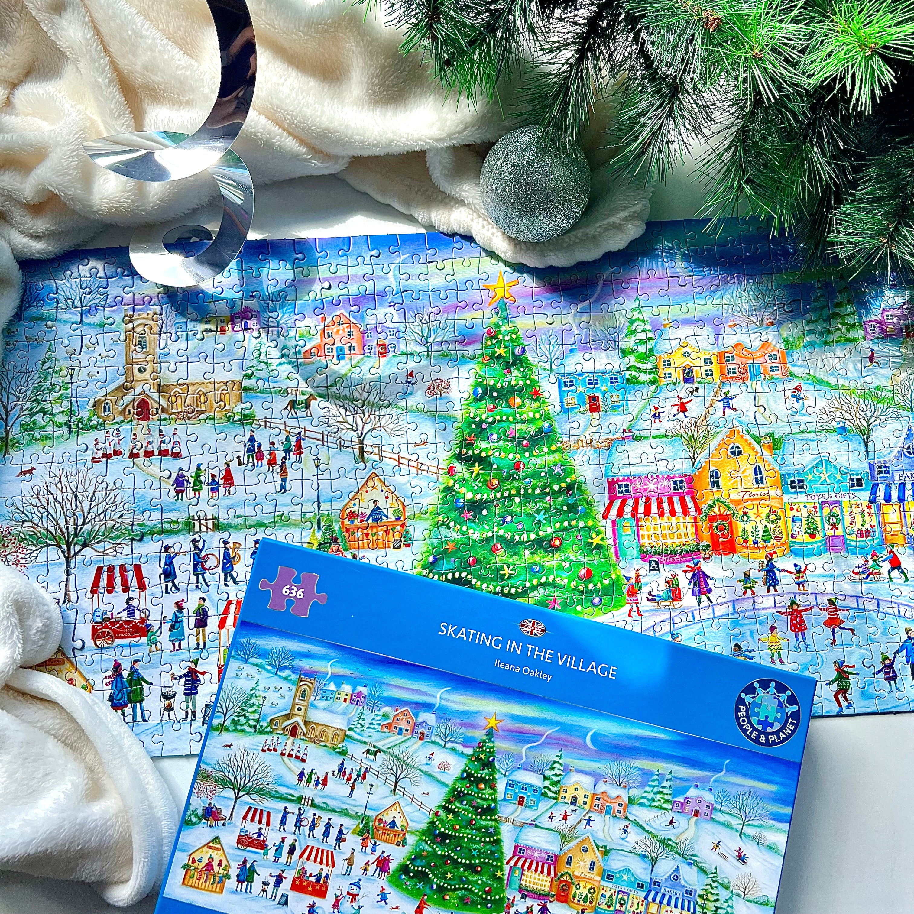 A colourful winter village jigsaw puzzle and its blue packaging box, styled with a plush cream blanket, a silver ornament, and evergreen branches.