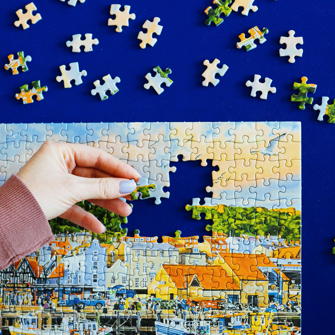 A hand is placing a jigsaw puzzle piece onto a colorful coastal town puzzle, with many loose puzzle pieces scattered on a dark blue background.