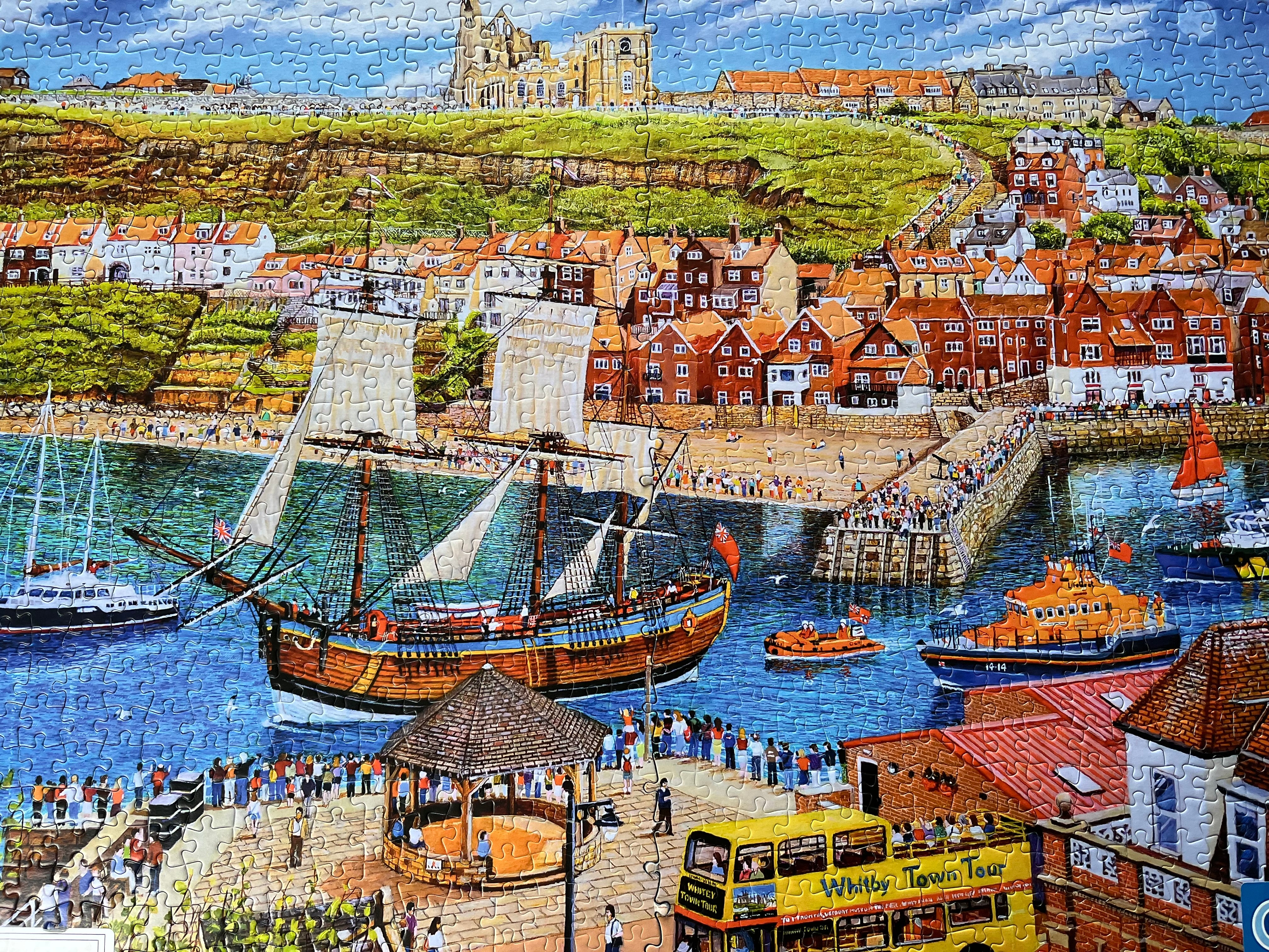 A completed jigsaw puzzle depicting a colourful coastal harbour scene with a large sailing ship, smaller boats, stone walkways, red-roofed buildings, and a hillside town under a blue sky.