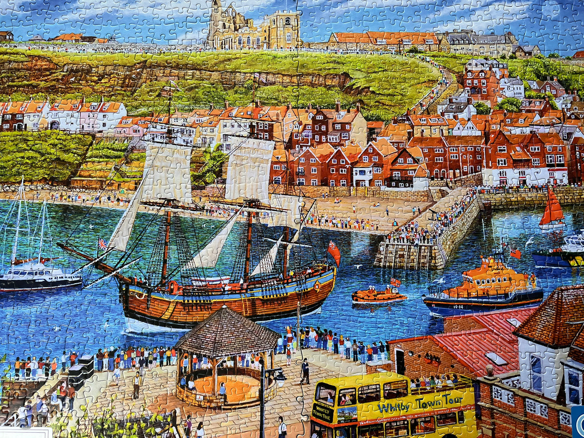 A completed jigsaw puzzle depicting a colourful coastal harbour scene with a large sailing ship, smaller boats, stone walkways, red-roofed buildings, and a hillside town under a blue sky.