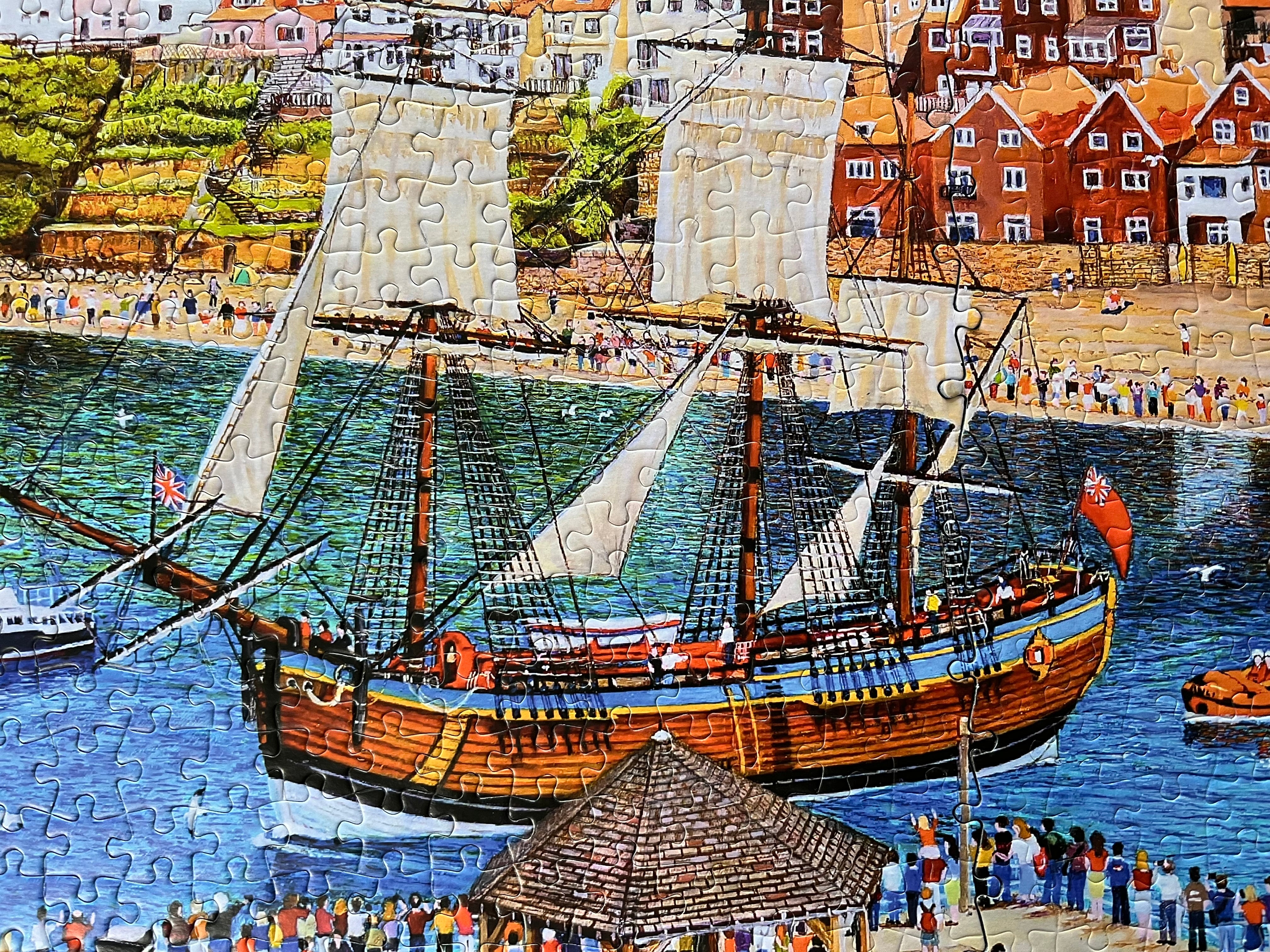 A colourful jigsaw puzzle depicting a coastal harbour scene with a large wooden sailing ship, blue water, beachgoers along the shore, and red-roofed buildings.