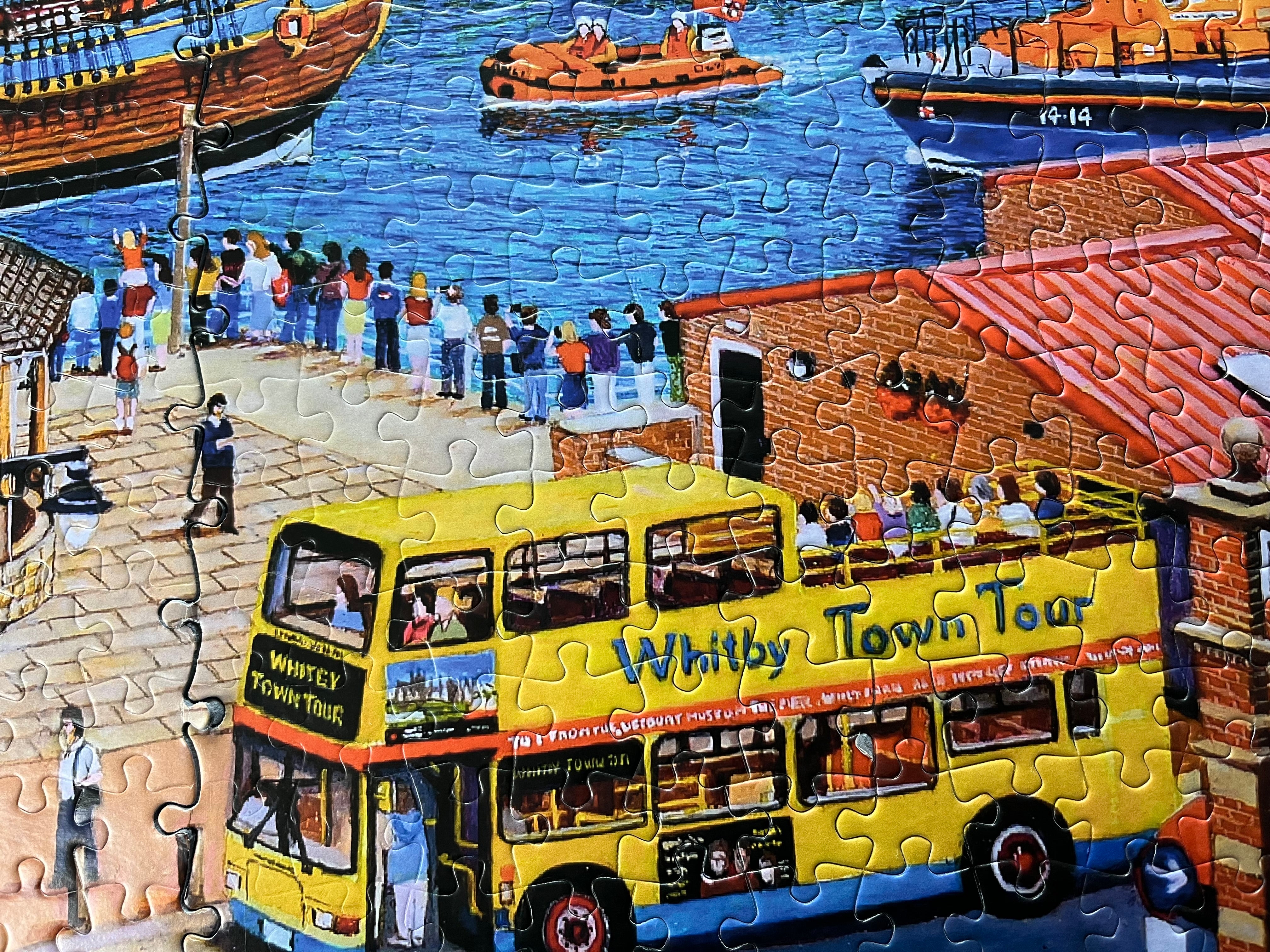A completed jigsaw puzzle showing a colourful coastal town scene with a yellow double-decker bus labelled 'Whitby Town Tour,' boats on blue water, pedestrians, and brick buildings.