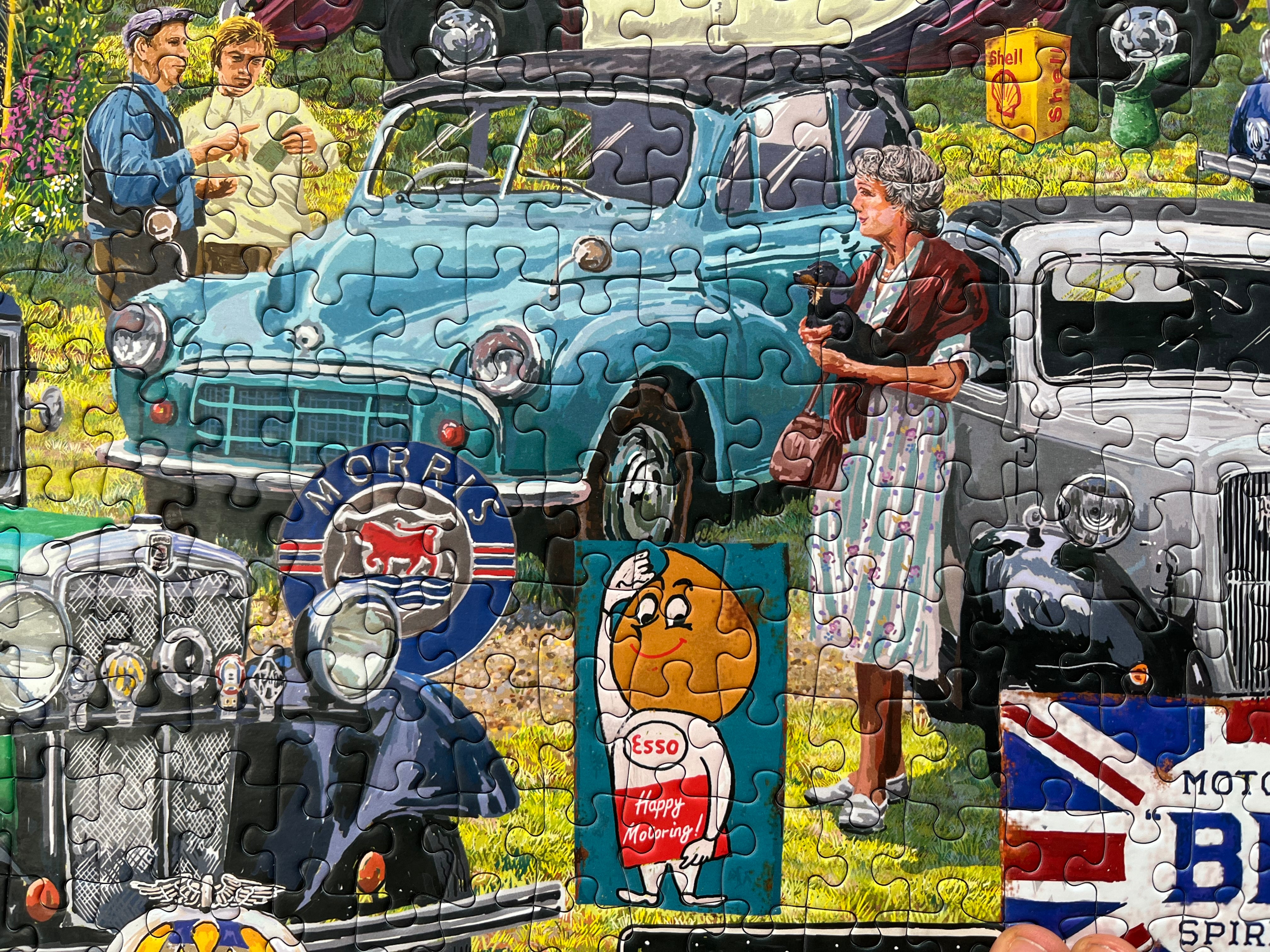 A completed jigsaw puzzle showing a vintage roadside scene with a teal car, a woman in a striped dress, other classic cars, and retro signs including an Esso logo.