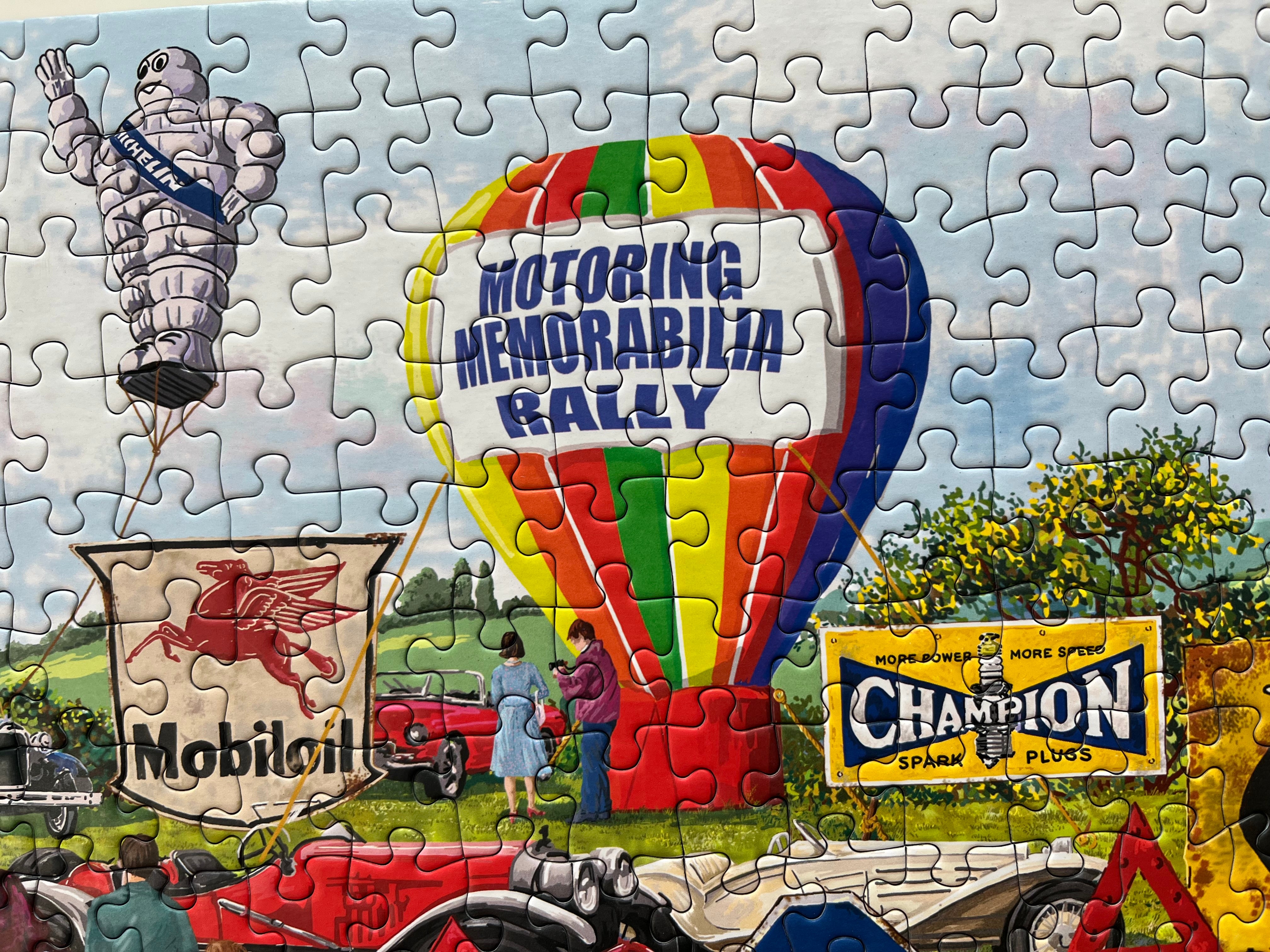 A completed jigsaw puzzle showing a vintage racing and advertising scene with a large hot air balloon reading 'Motoring Memorabilia Rally,' a Mobil 1 sign, a Champion Spark Plugs sign, and classic cars on a grassy field.