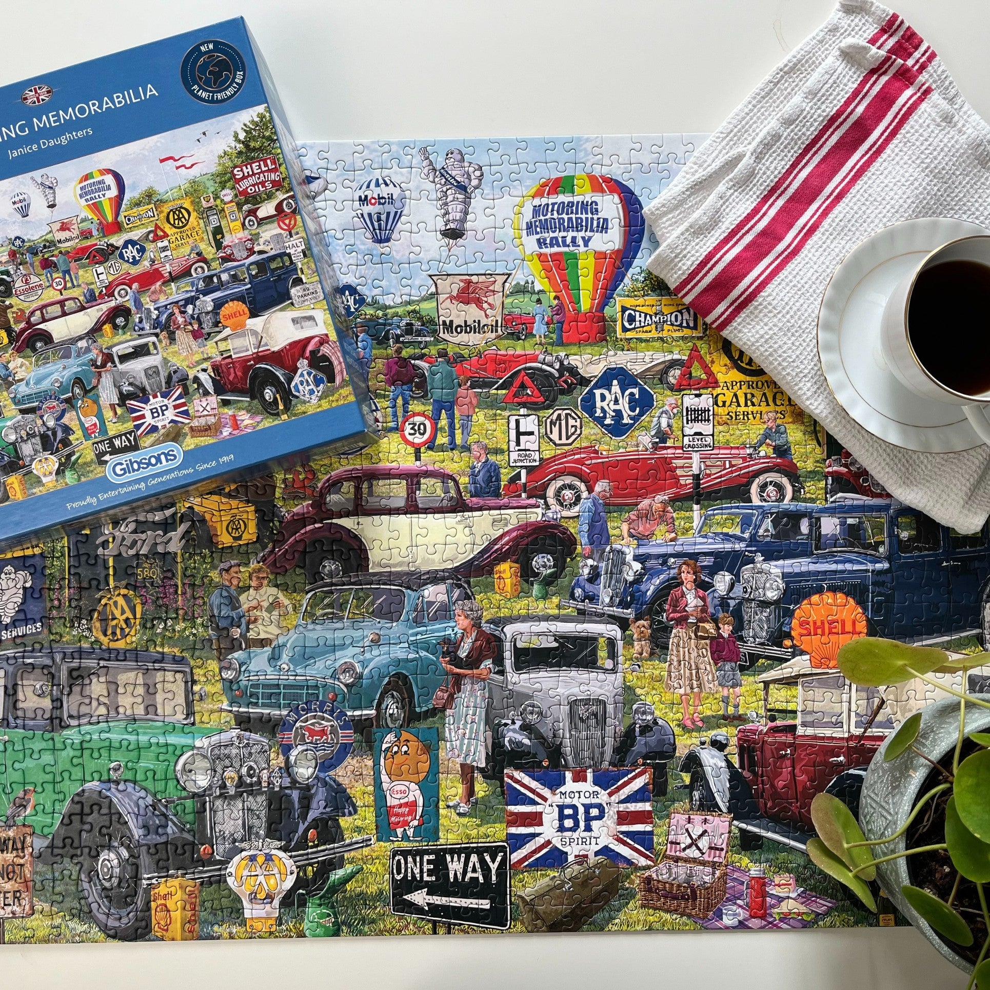 A colourful vintage car-themed jigsaw puzzle and its blue packaging box are arranged on a white tabletop, alongside a striped tea towel, a cup of black coffee, and a potted plant.
