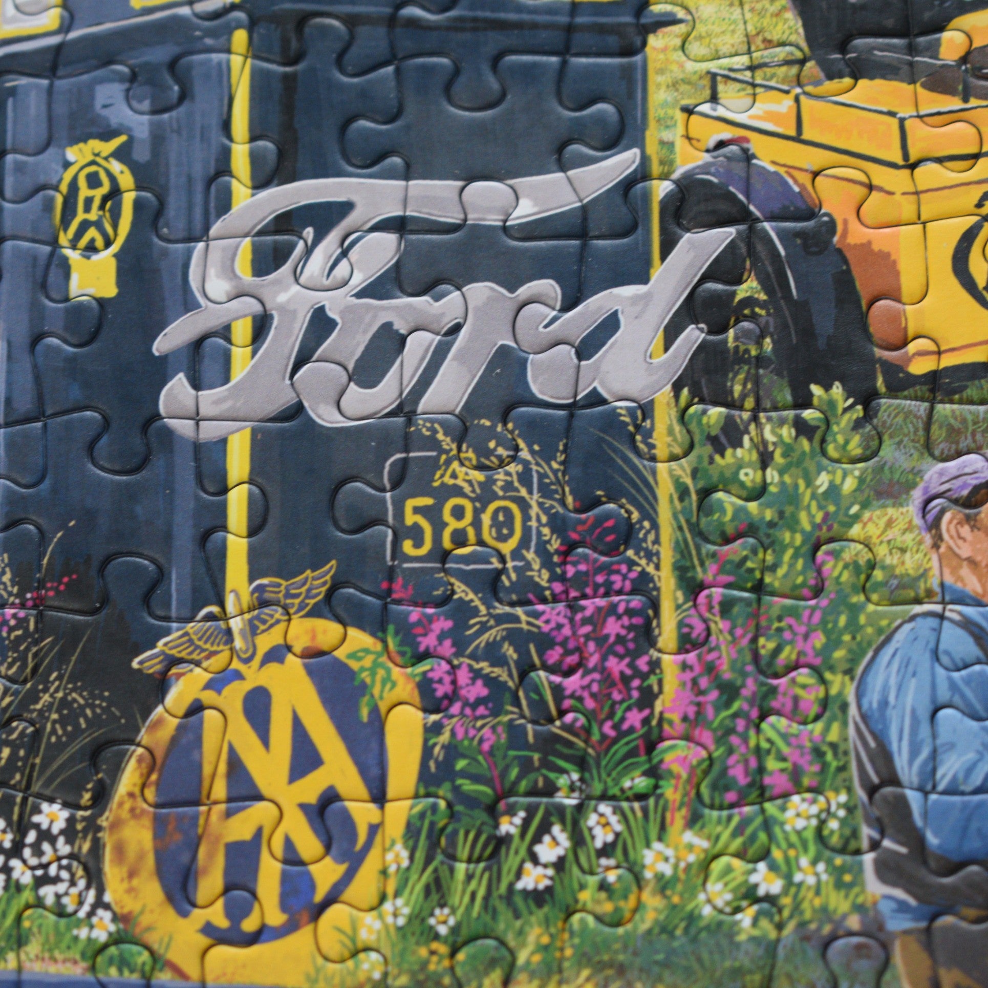 A close-up of a completed jigsaw puzzle showing a stylized Ford-themed design with the Ford script logo, yellow number 580, floral artwork, and part of a person in the lower right corner.