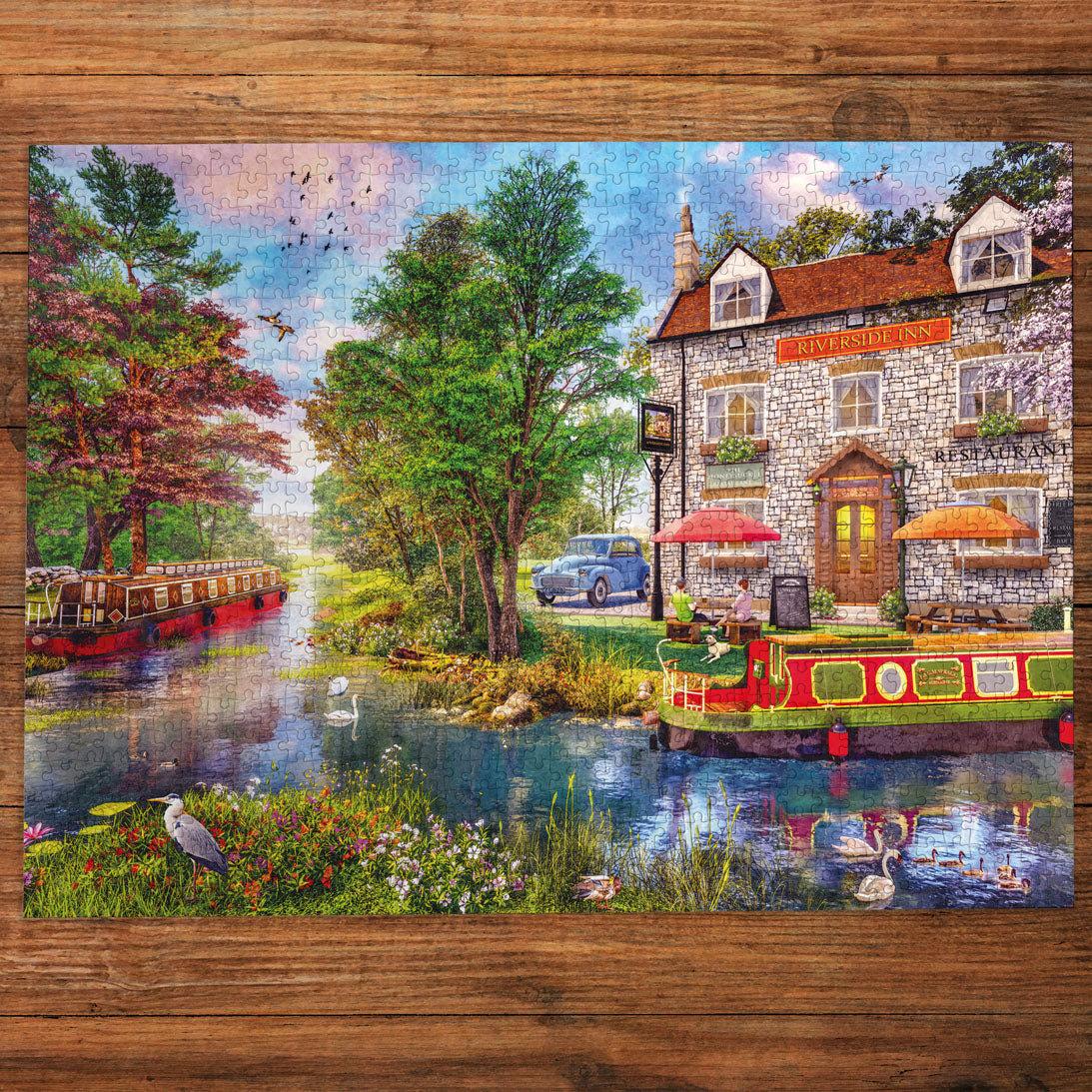 A completed jigsaw puzzle depicting a picturesque stone pub by a river, with boats, trees, ducks, and a blue car, displayed on a rustic wooden surface.