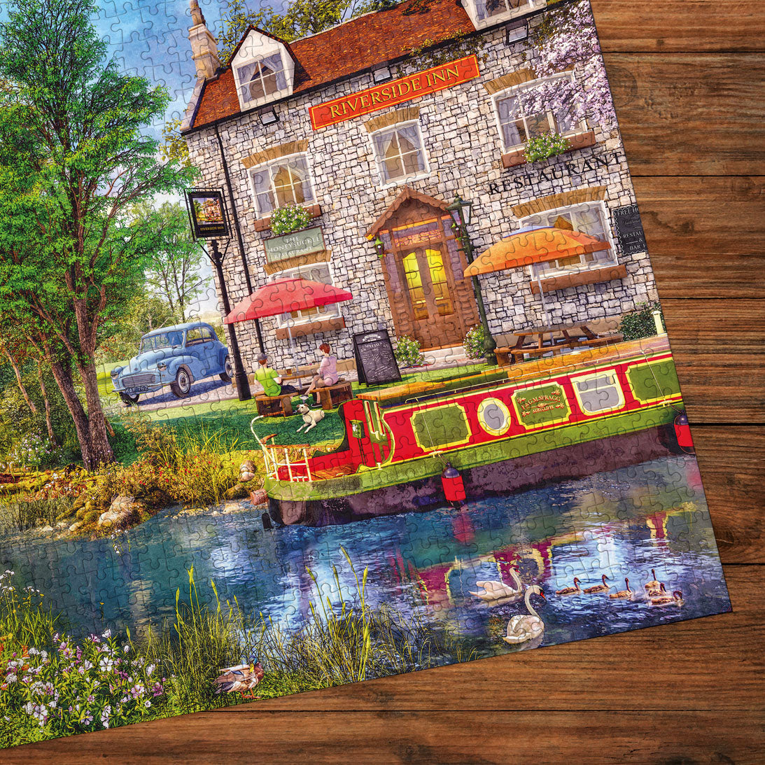 A completed jigsaw puzzle depicting a picturesque stone pub named Riverside Inn by a canal, with a red and green boat, ducks, swans, trees, and a blue vintage car, shown placed on a wooden surface.