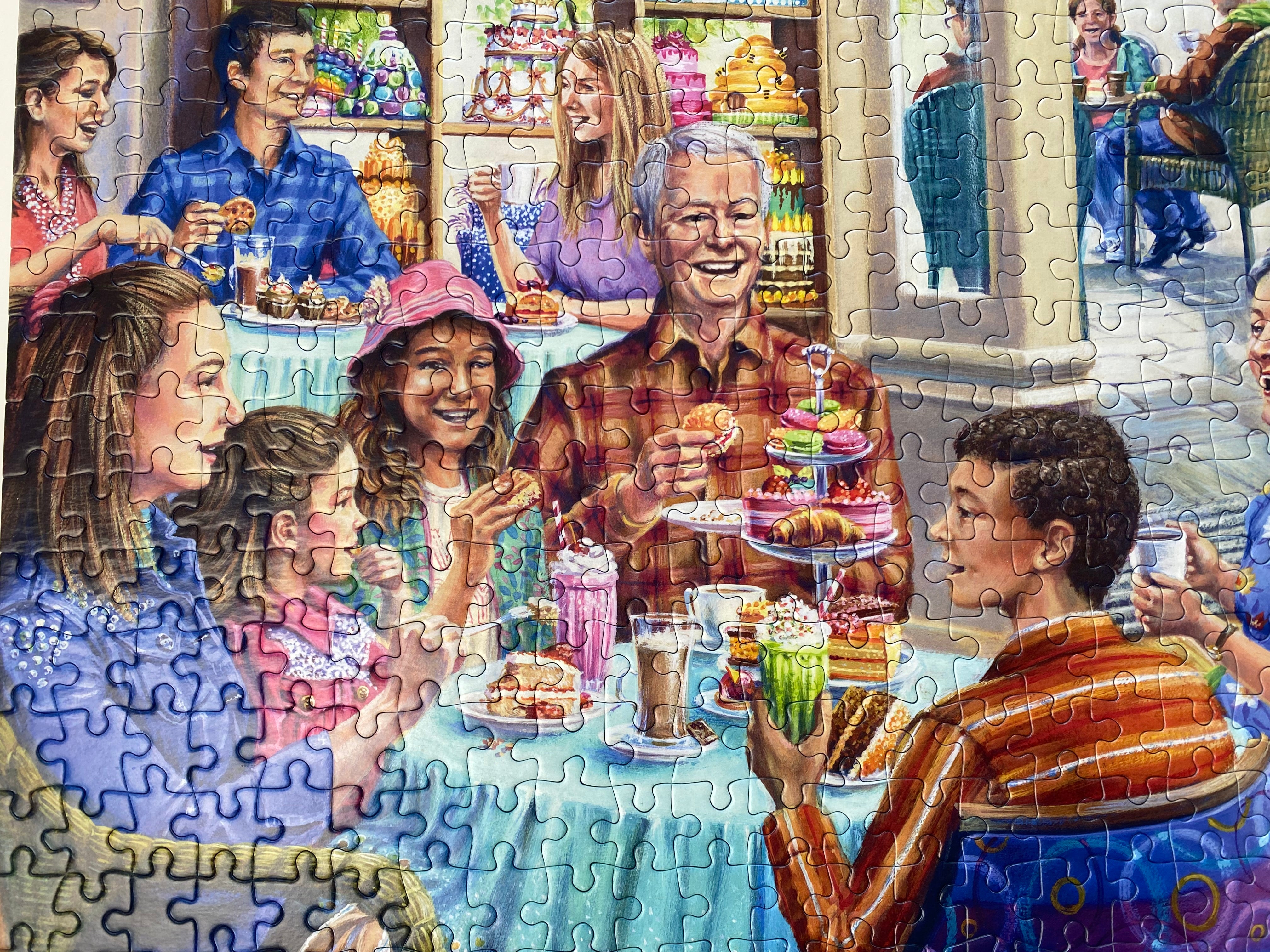 A completed jigsaw puzzle showing a colourful painted scene of a family and friends gathered around a table with pastries, drinks, and baked goods in a bakery or cafe setting.