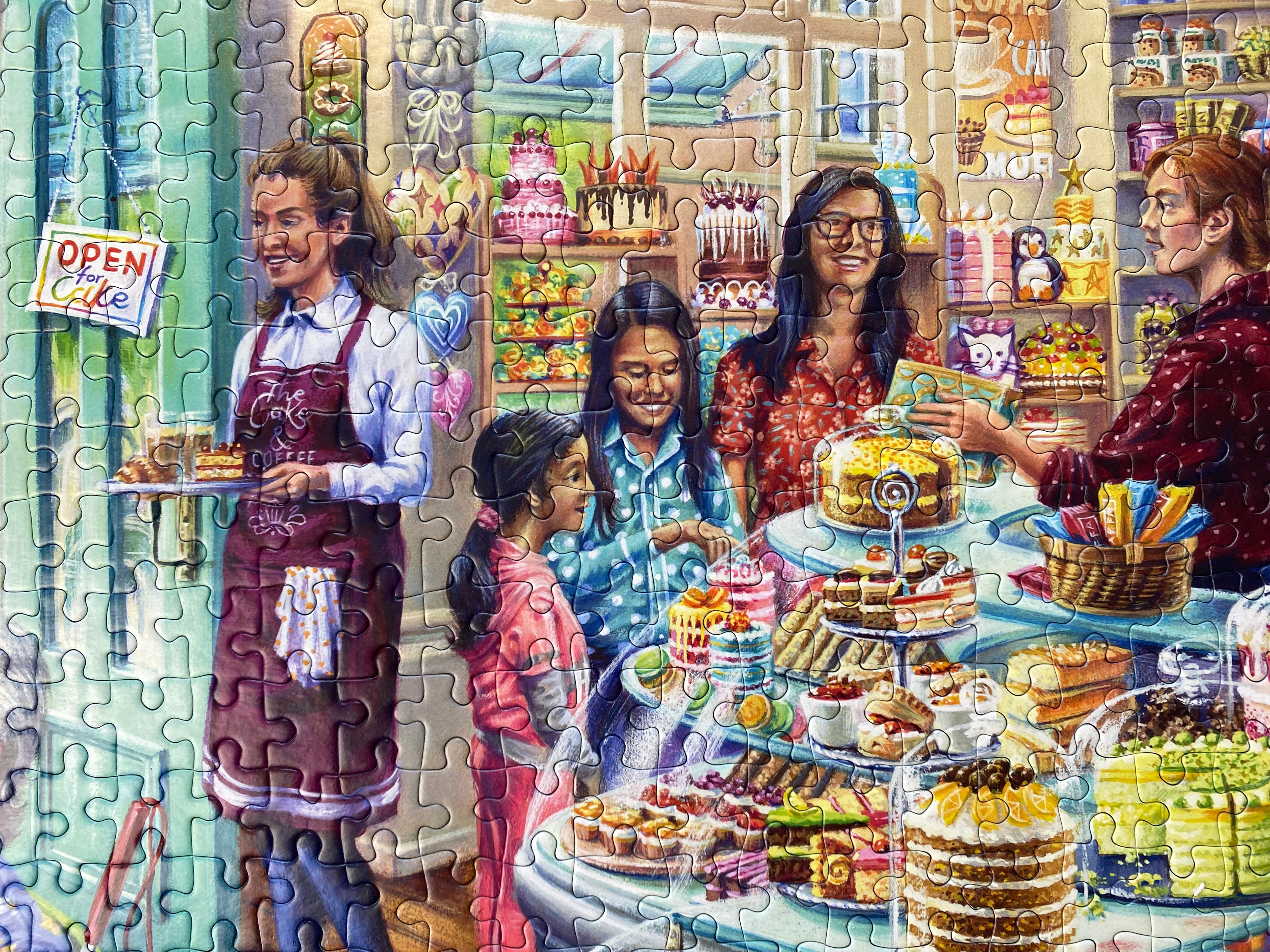 A completed jigsaw puzzle showing a colourful bakery or cafe interior with several people gathered around a counter filled with cakes, pastries, and desserts. The puzzle pieces are clearly visible across the entire scene.