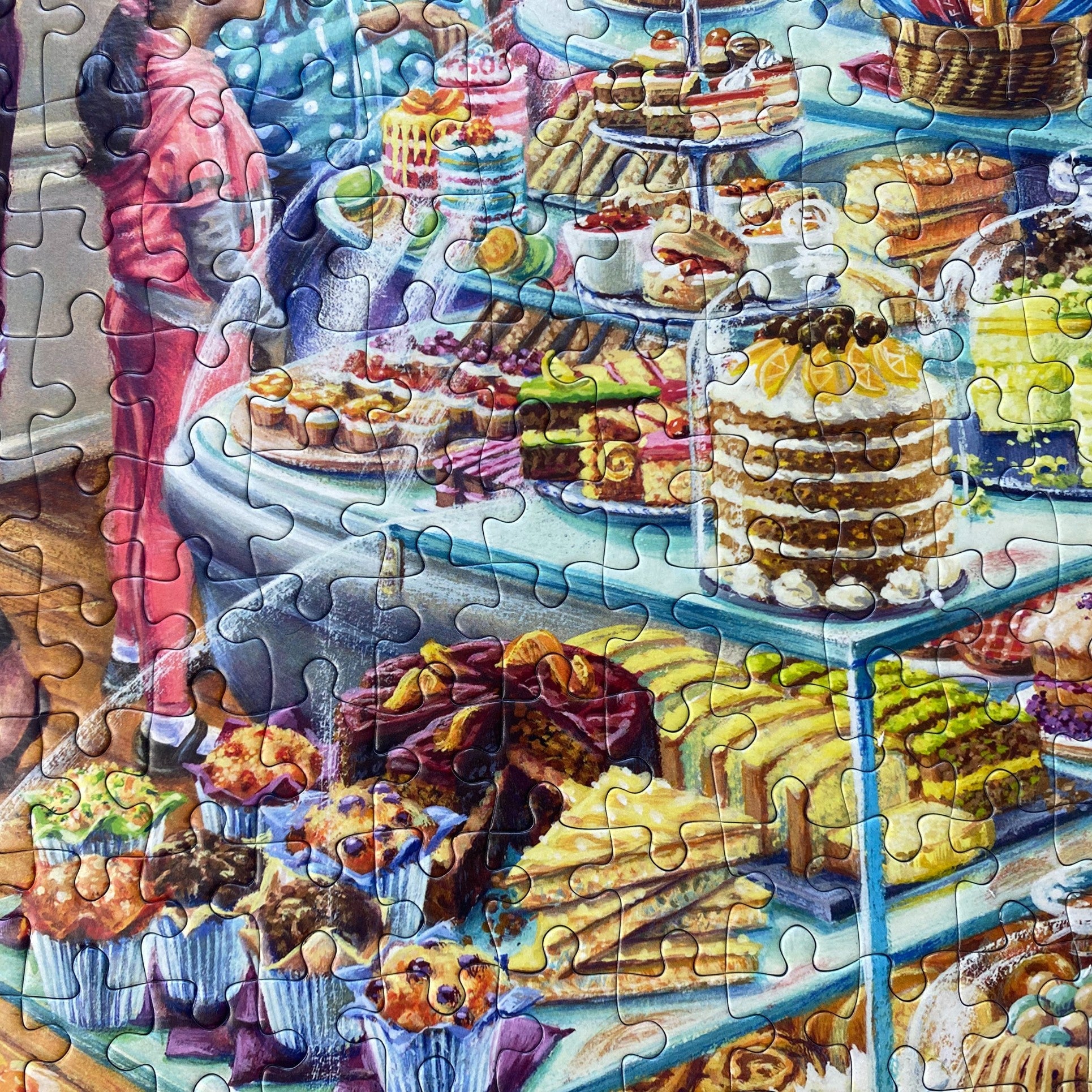 A colourful jigsaw puzzle showing a detailed bakery and dessert display with cakes, pastries, cupcakes, and a person in a pink outfit, with puzzle piece cutlines visible across the surface.