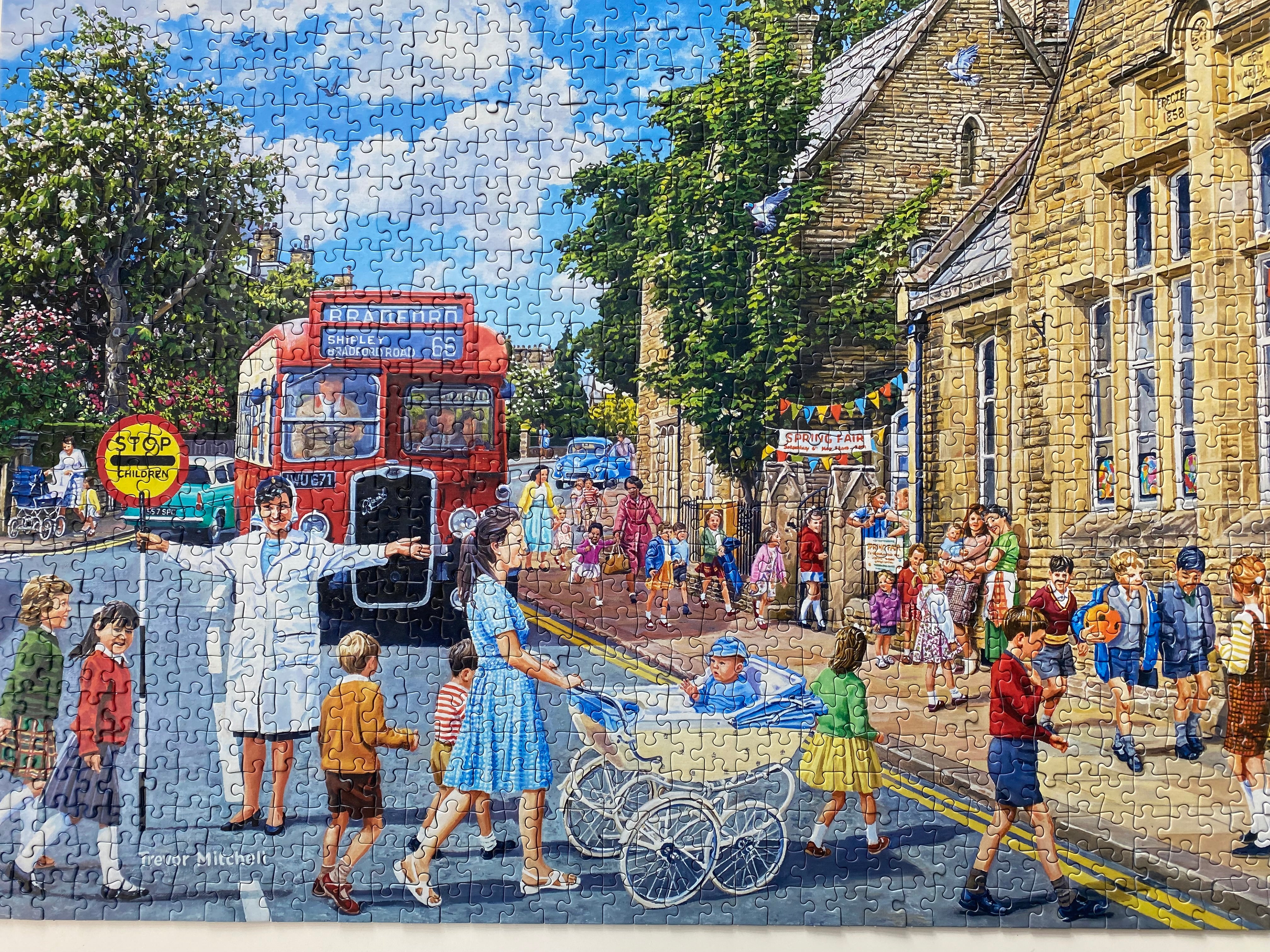 A completed jigsaw puzzle showing a colourful village street scene with children, a red bus, trees, and a stone building.