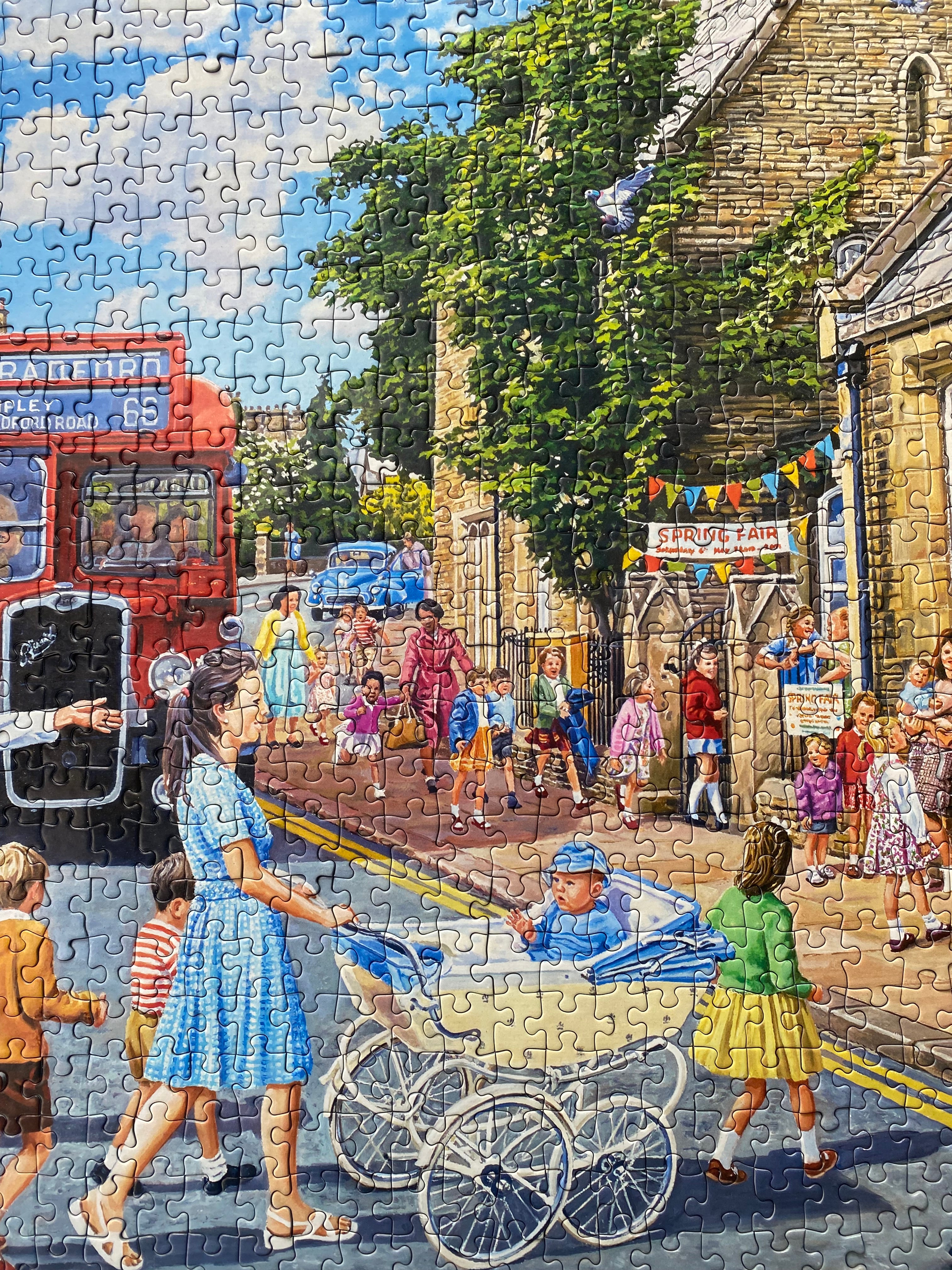 A completed jigsaw puzzle showing a colourful village street scene with children, a red bus, trees, and a stone building, photographed straight on.