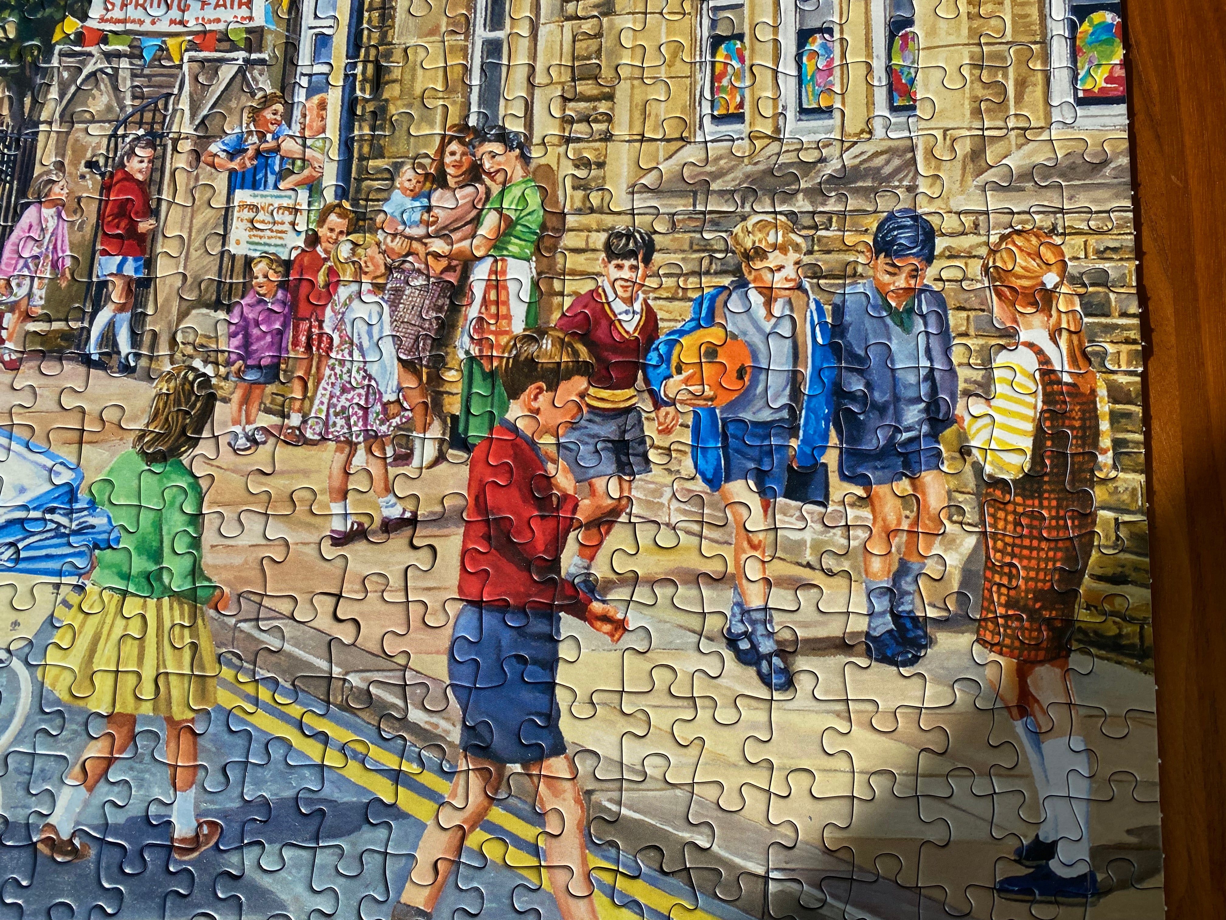 A completed jigsaw puzzle showing a colourful illustration of children playing on a street in front of a stone building, photographed close up against a wooden surface.