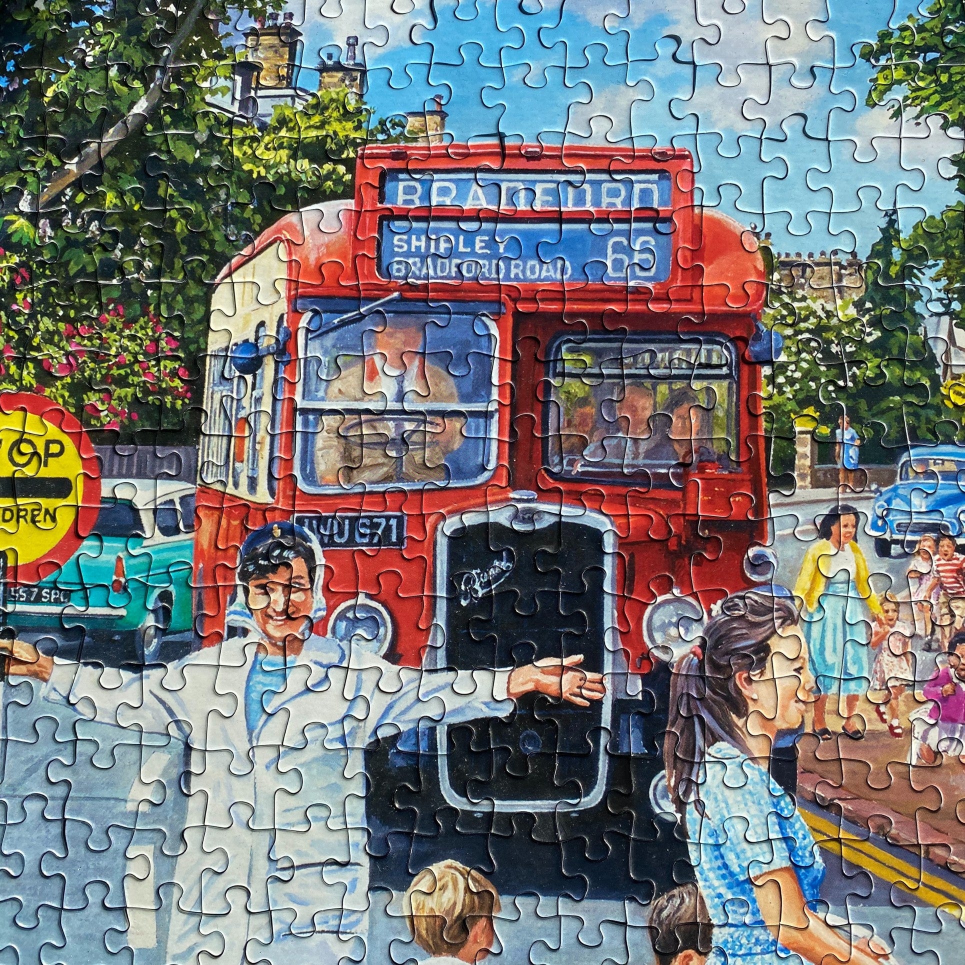 A completed jigsaw puzzle showing a colorful painted street scene with a red double-decker bus, trees, pedestrians, and vintage cars. The puzzle pieces are visible across the entire image.