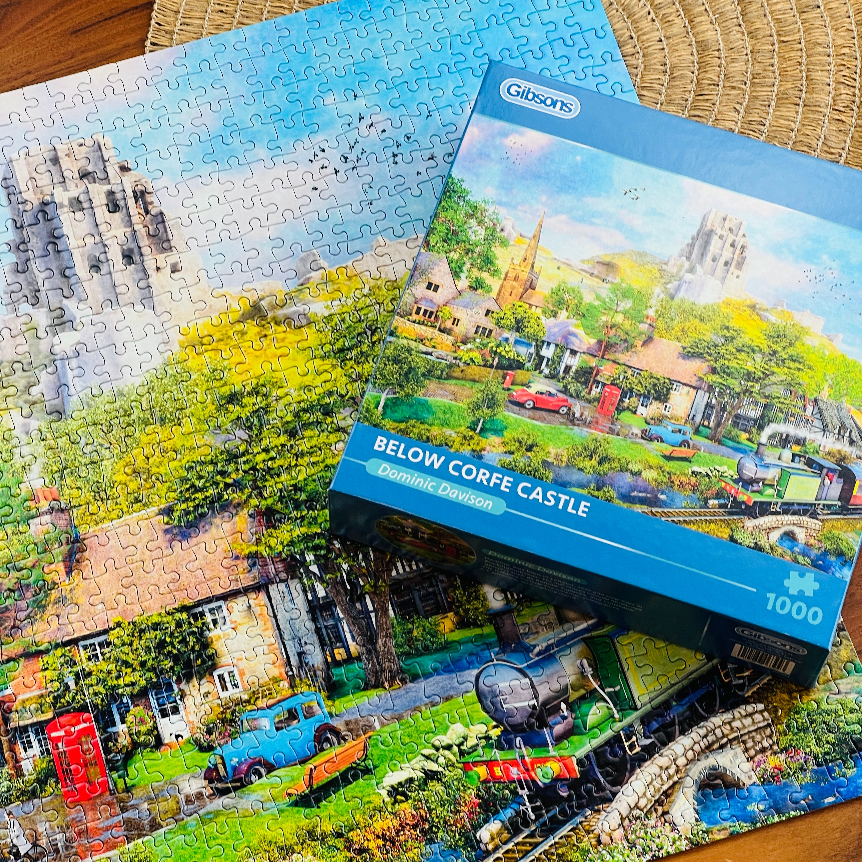 A 1000-piece jigsaw puzzle and its blue retail box from Gibsons, featuring a coloruful village scene with a castle, trees, houses, and a train, arranged on a woven mat over a wooden surface.