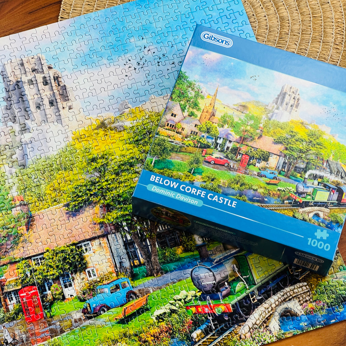 A 1000-piece jigsaw puzzle and its blue retail box from Gibsons, featuring a coloruful village scene with a castle, trees, houses, and a train, arranged on a woven mat over a wooden surface.
