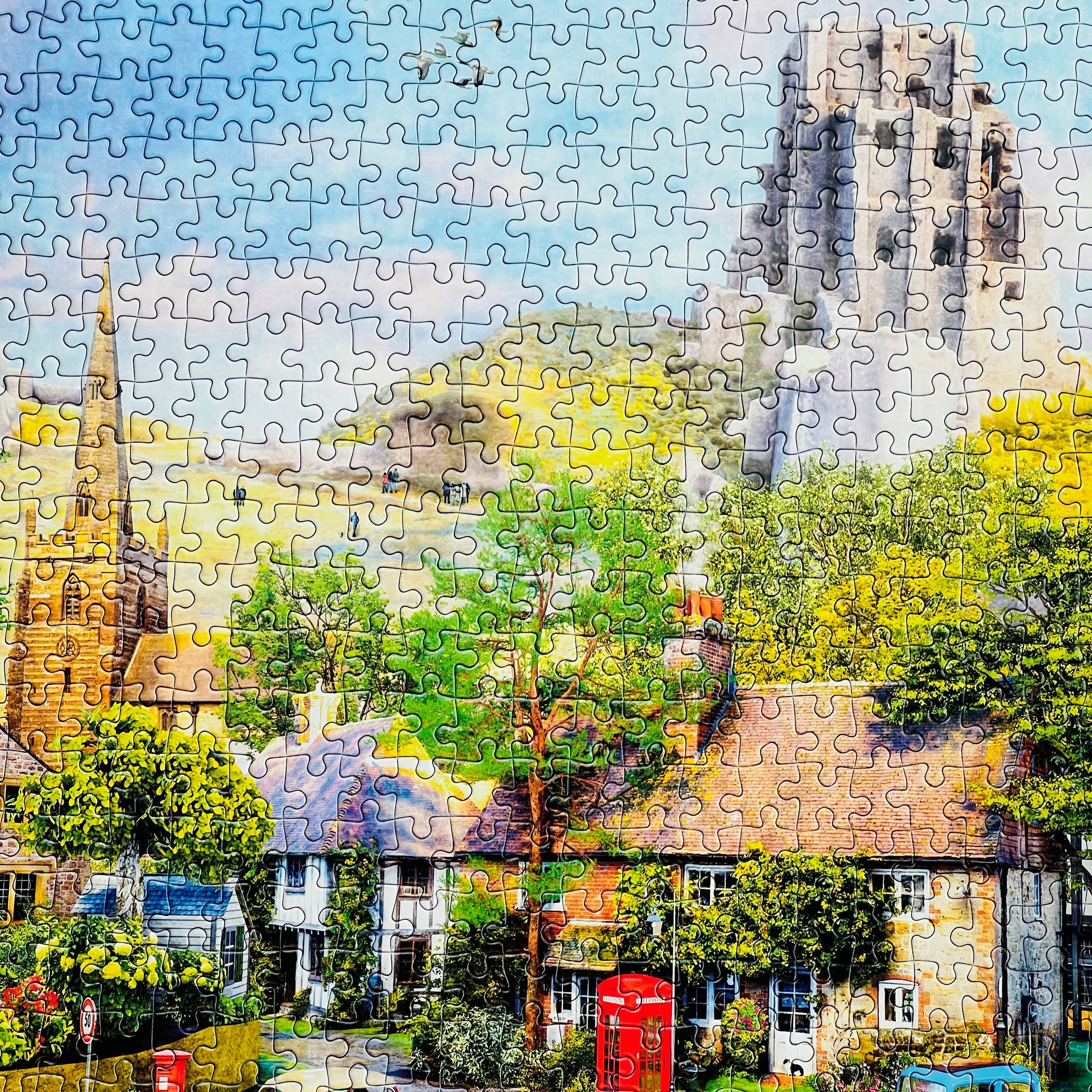 A completed jigsaw puzzle showing a colourful village scene with cottages, trees, a red telephone booth, and a large stone tower under a blue sky.