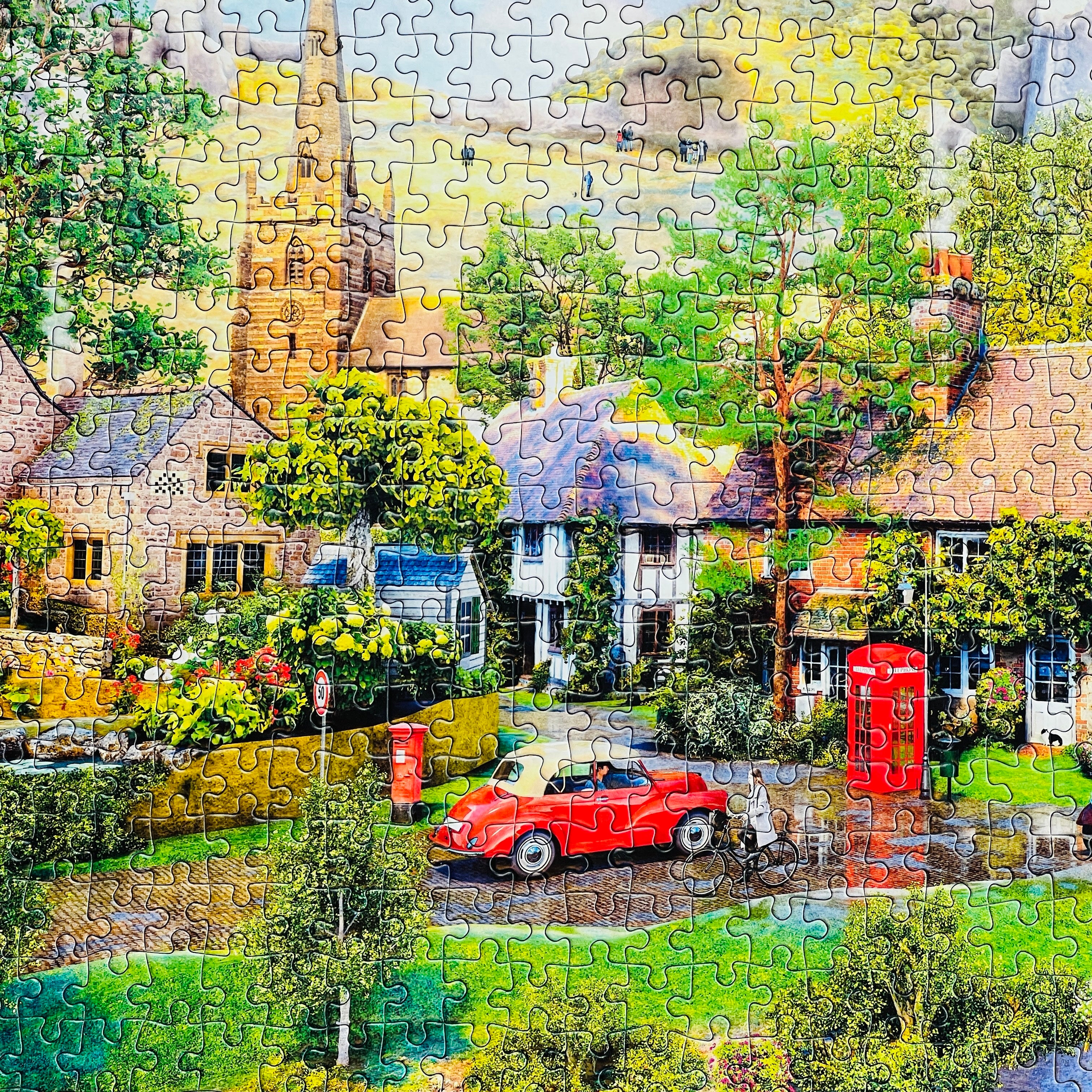A completed jigsaw puzzle showing a colourful village street scene with cottages, trees, a red car, a red telephone booth, and a church tower in the background.