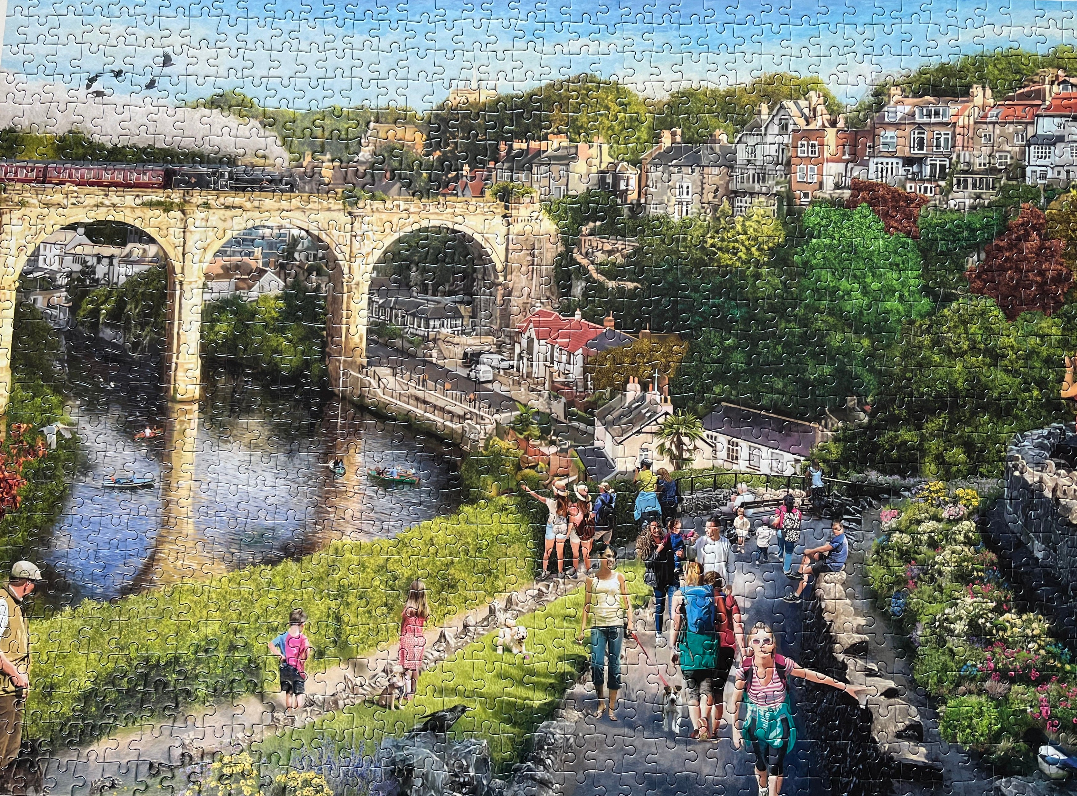A completed jigsaw puzzle showing a scenic landscape with a large stone bridge over a river, trees, buildings, and groups of people walking along a path. The puzzle pieces are clearly visible across the entire image.
