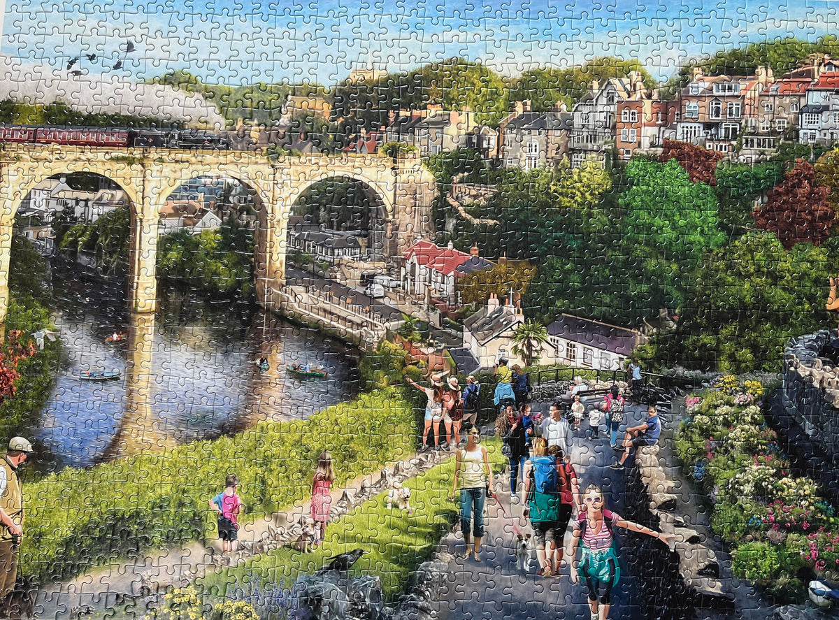 A completed jigsaw puzzle showing a scenic landscape with a large stone bridge over a river, trees, buildings, and groups of people walking along a path. The puzzle pieces are clearly visible across the entire image.