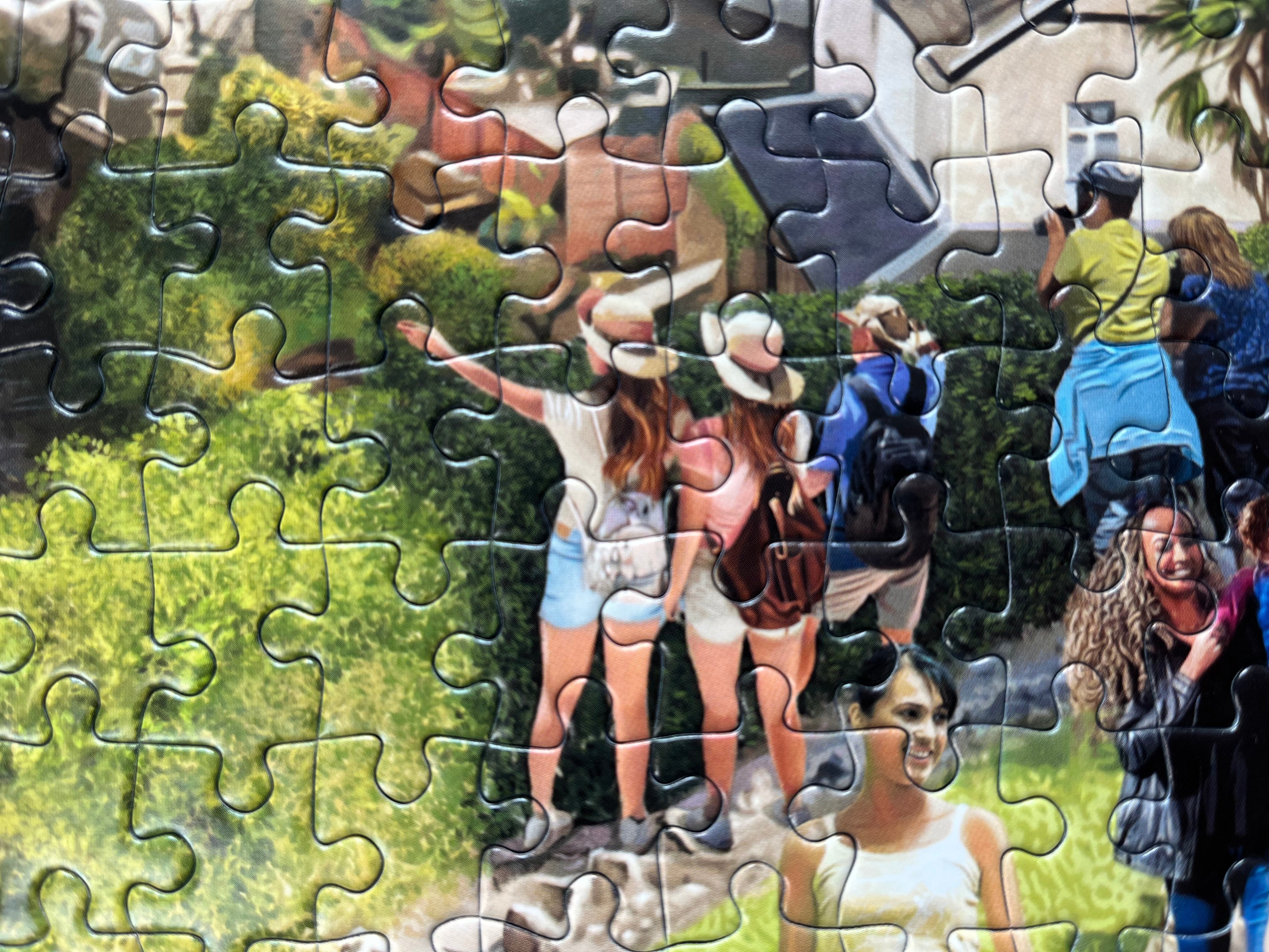 A completed jigsaw puzzle showing a colorful outdoor scene with several people standing on rocks in a grassy area, with puzzle pieces still visible across the entire surface.